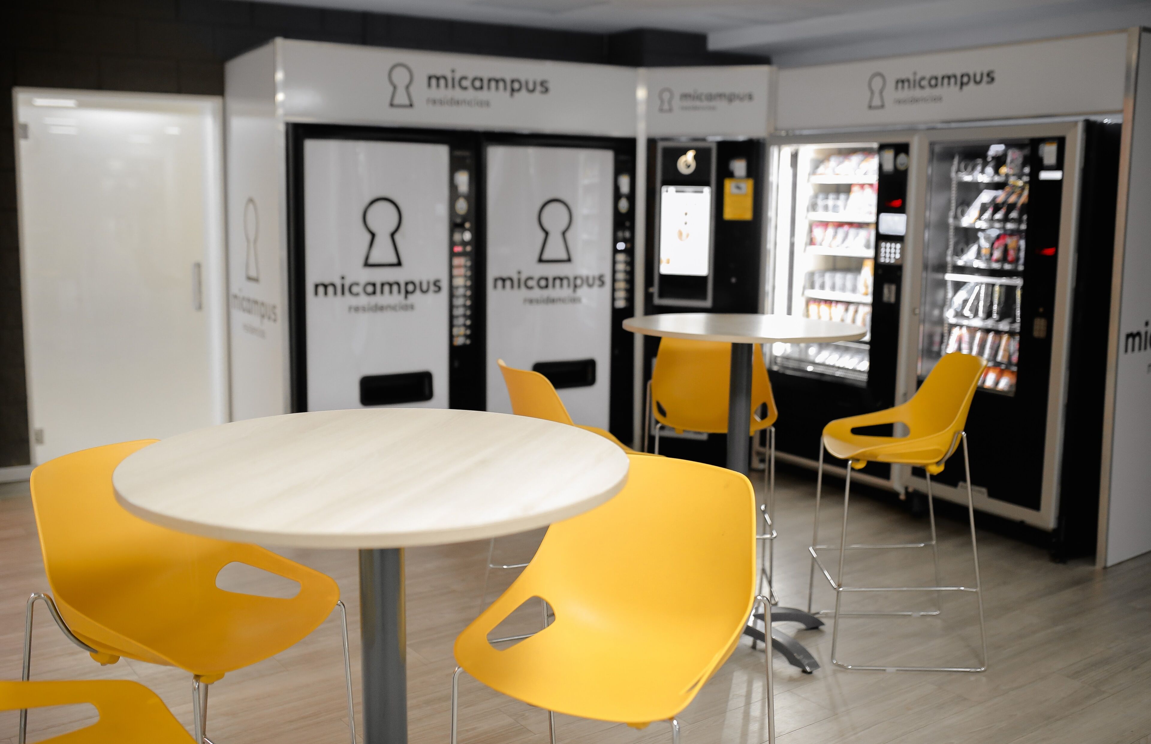 A room with a round table and four yellow chairs, vending machines labeled 'micampus residences', and a shoe display.