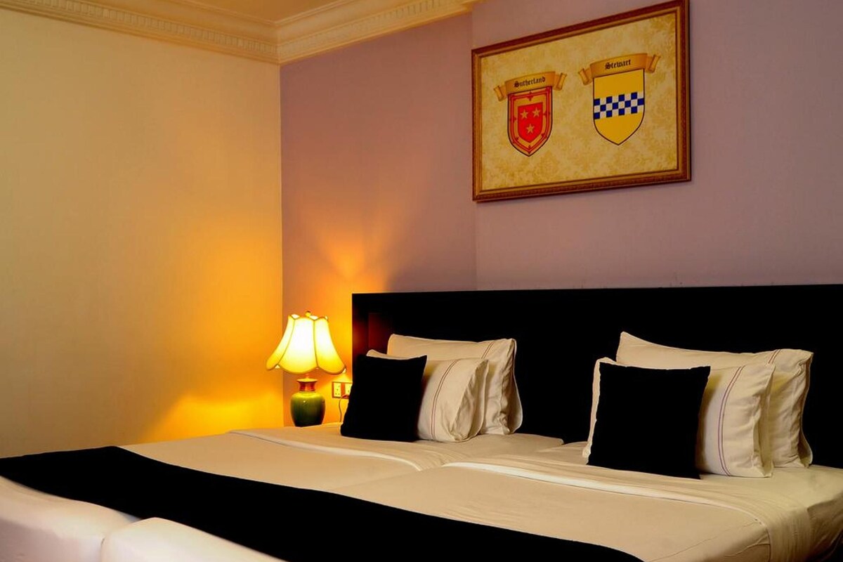 A hotel room with a large bed, two black pillows, a headboard, a lamp, and a framed coat of arms on the wall.
