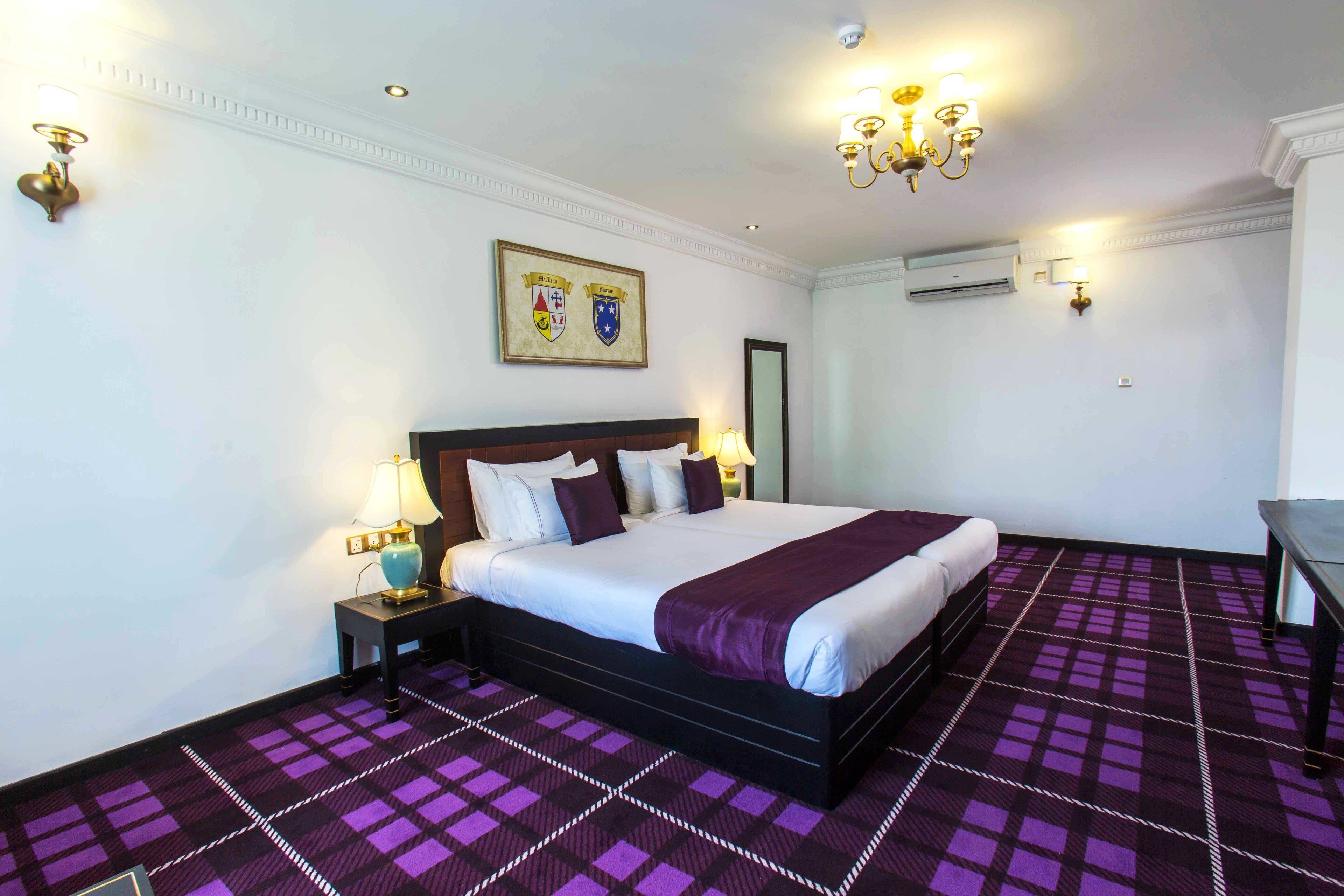 A hotel room with a large bed, a nightstand, a lamp, a wall-mounted air conditioner, and a patterned carpet.