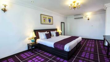 A hotel room with a large bed, a nightstand, a lamp, a wall-mounted air conditioner, and a patterned carpet.