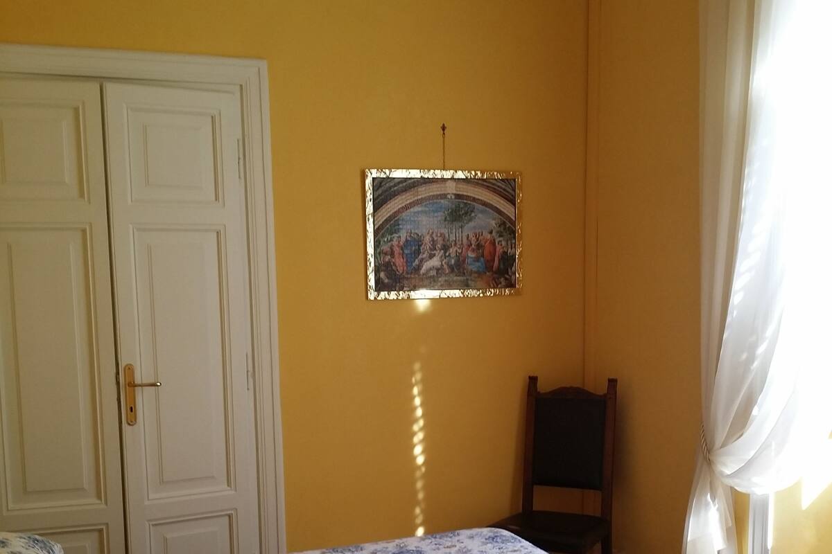 Ein Zimmer mit Bett, Tür, einem Bild im Rahmen an der Wand und einem Fenster mit Vorhängen.