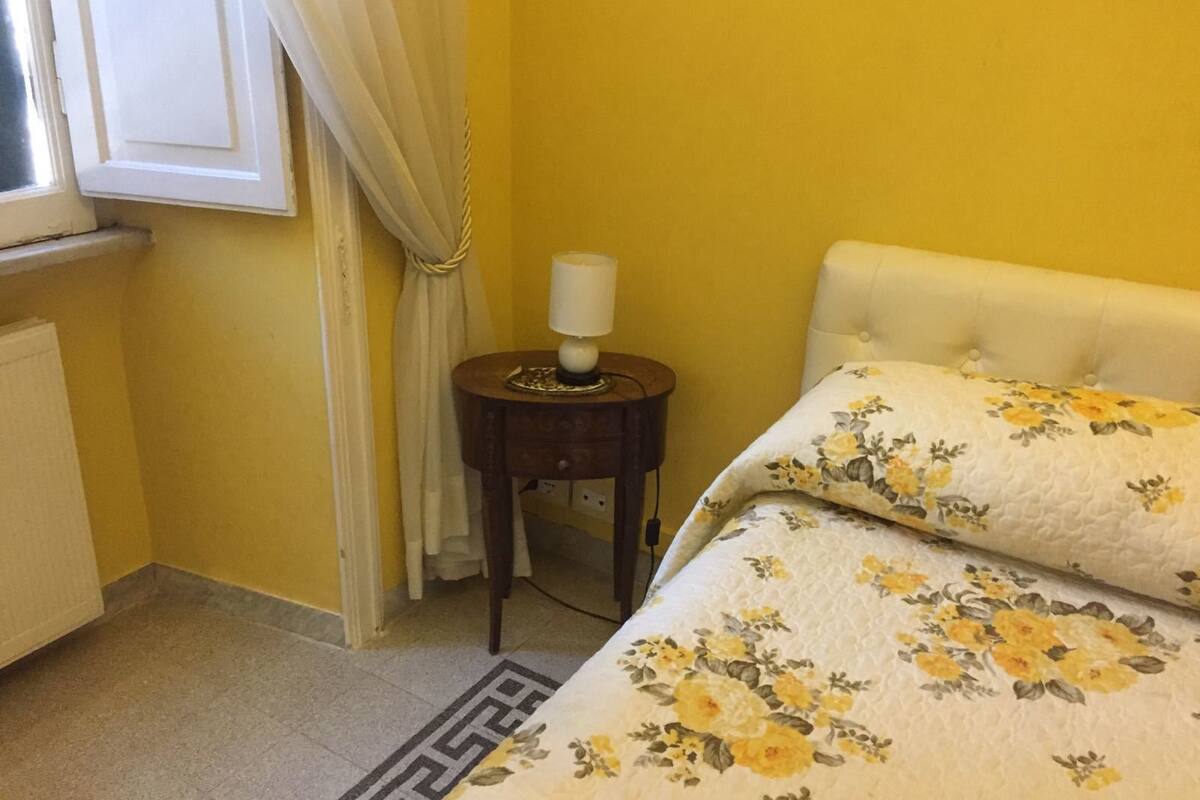 Ein Zimmer mit einem Bett, einem Nachttisch mit Lampe, einem Fenster mit Vorhängen und einer Heizung.