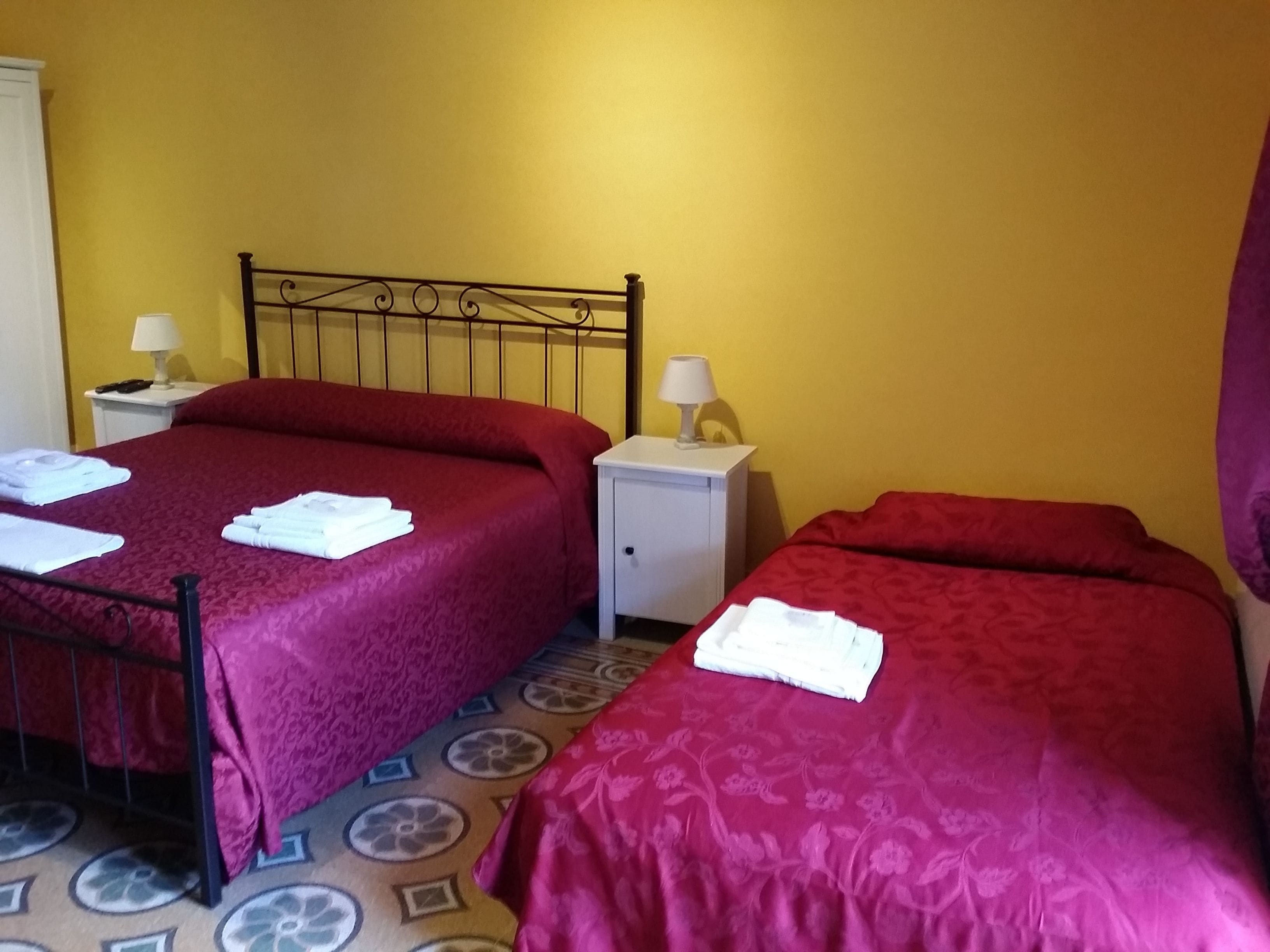 A hotel room with two beds, each with a red bedspread and white towels. There is a small bedside table with a lamp on each side of the beds.
