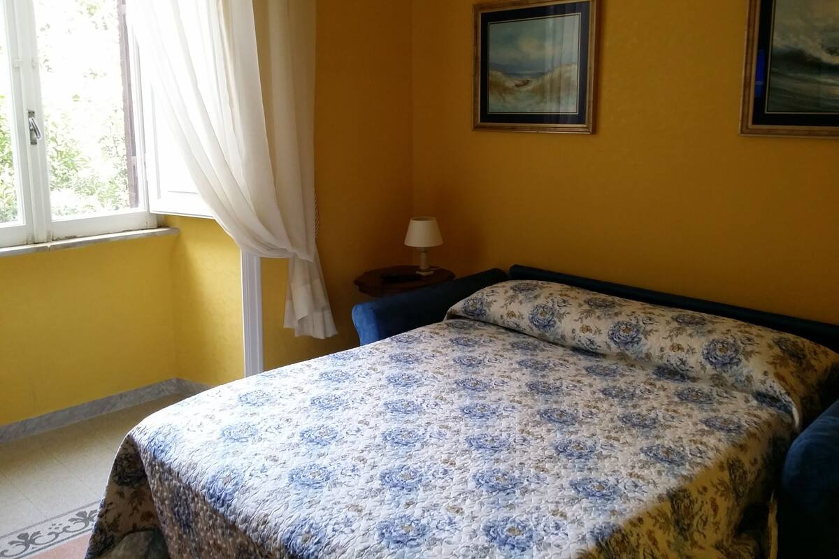 Ein Schlafzimmer mit Bett, einem Fenster mit Vorhängen, einem Nachttisch und zwei gerahmten Bildern an der Wand.