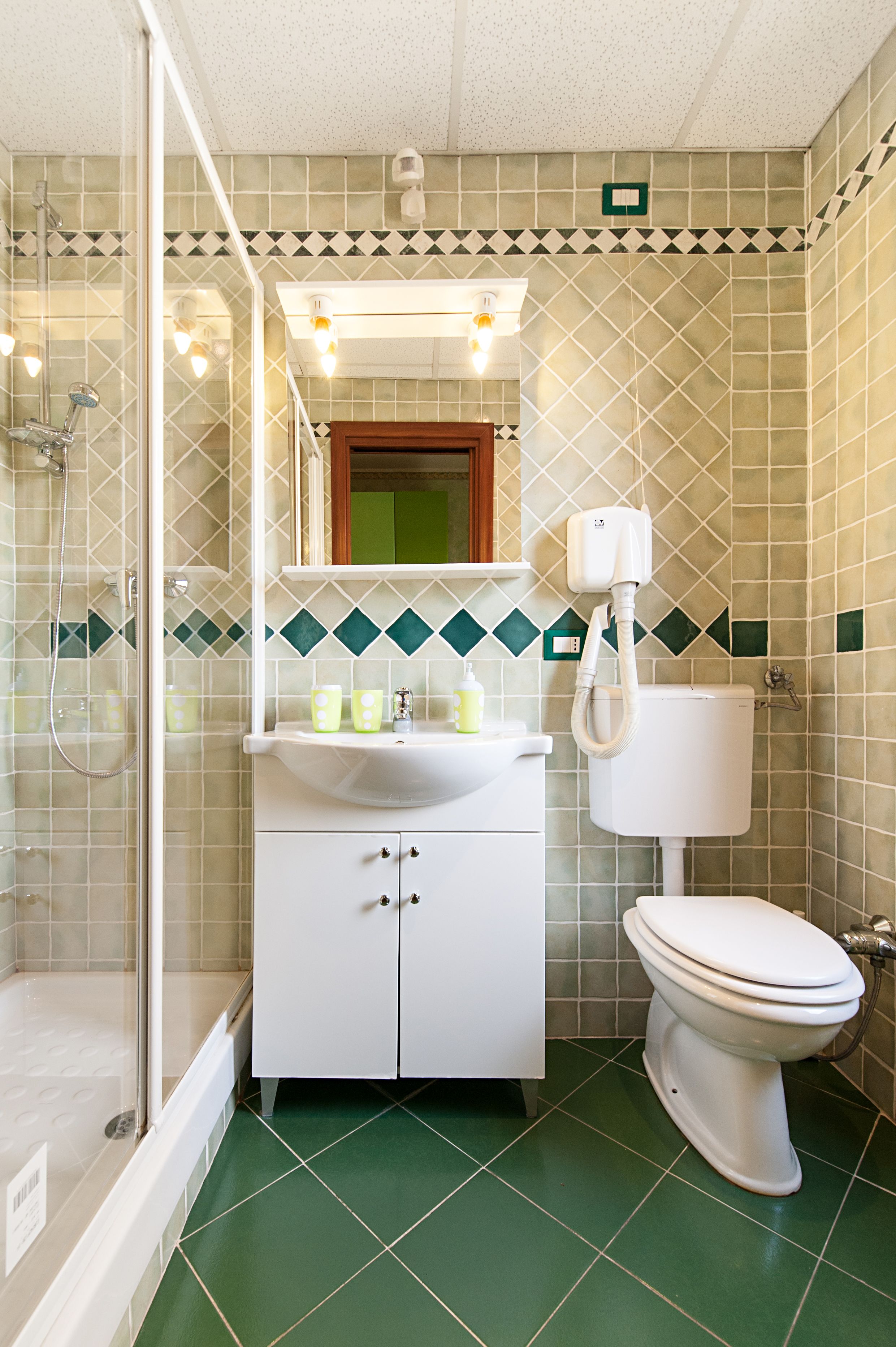 Un baño con ducha, inodoro y lavamanos, con azulejos verde y beige y un espejo sobre el lavamanos.