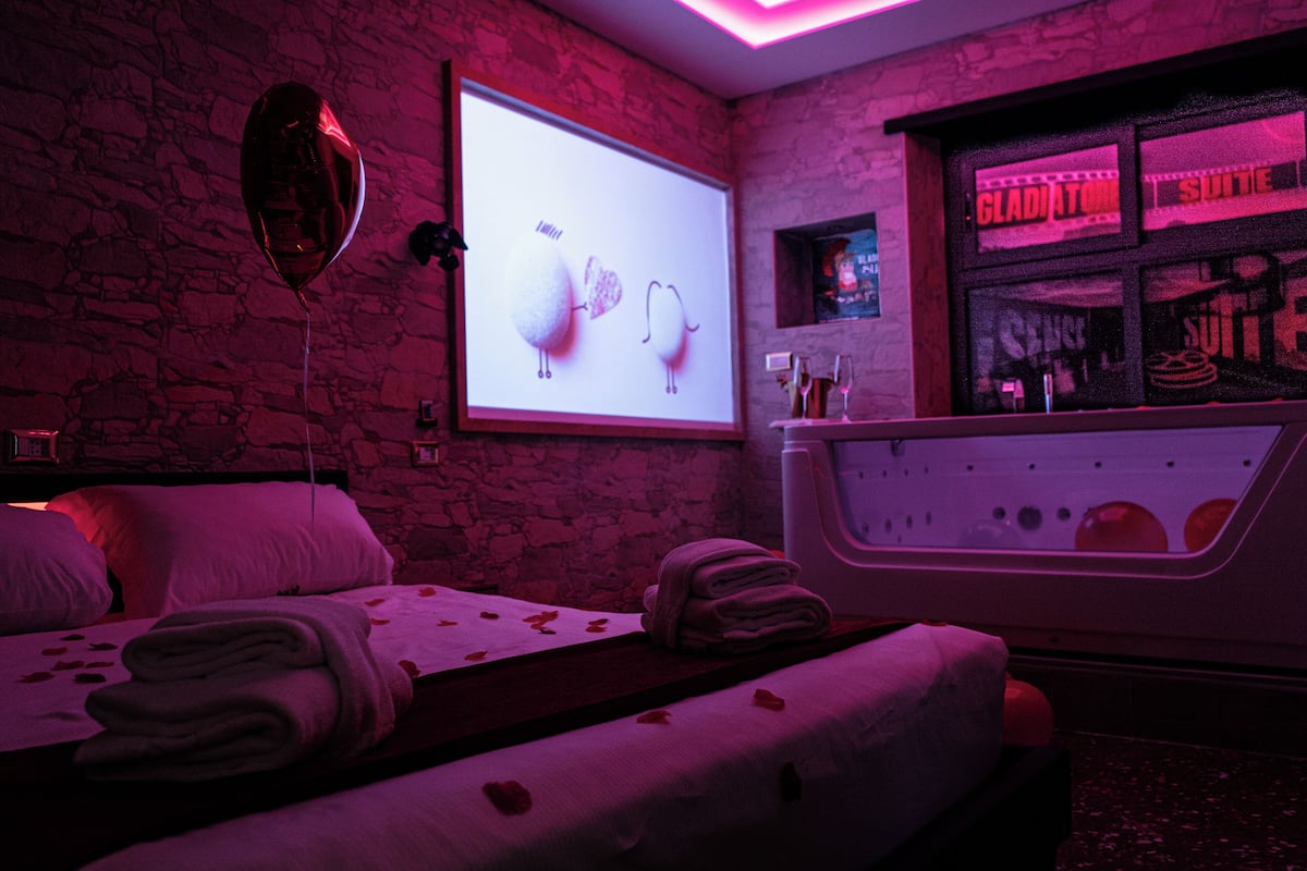 Una habitación con una cama, una silla de masaje, una pantalla mostrando una caricatura y una zona de bar con un letrero de neón.