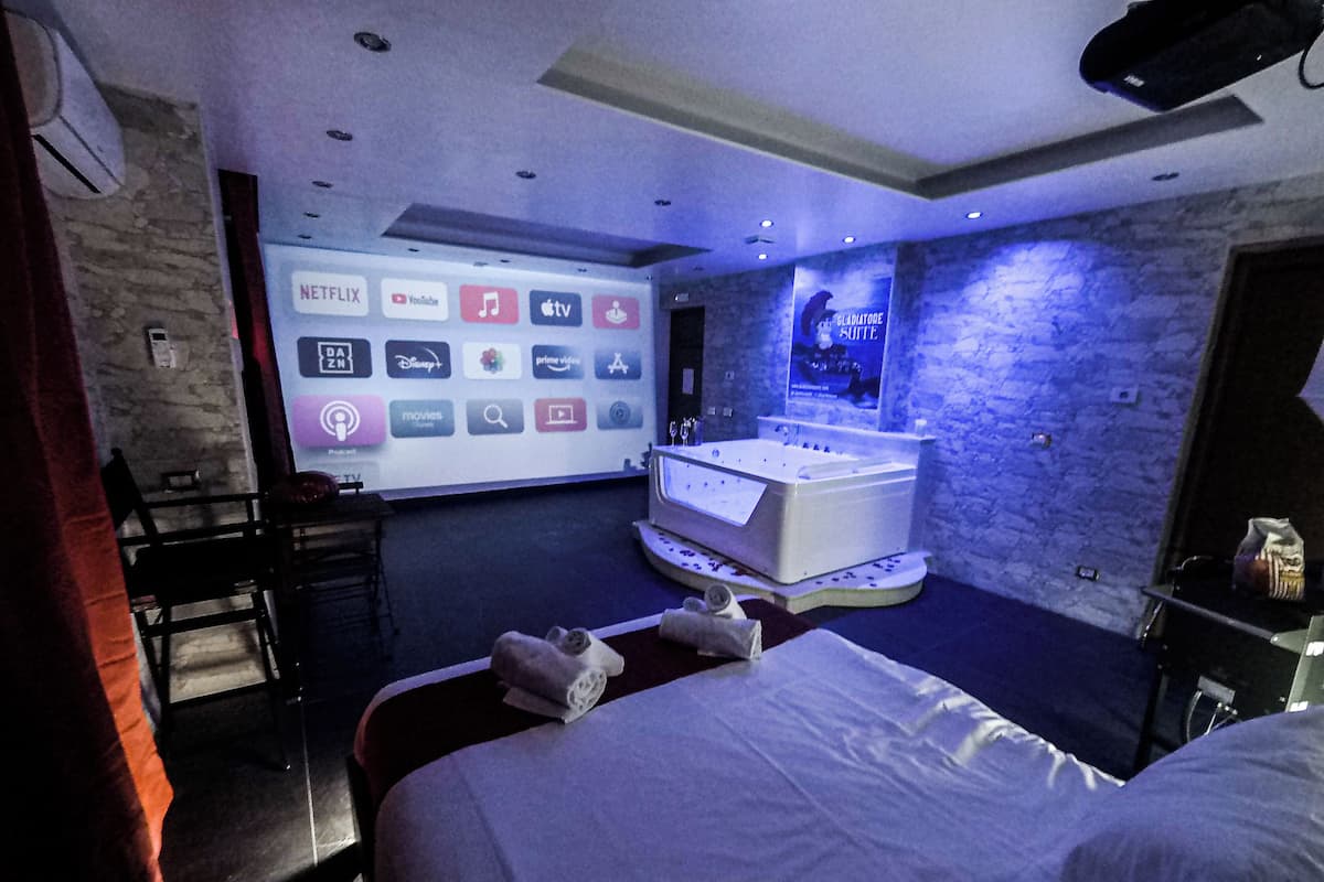 Habitación de hotel con cama, una pantalla grande que muestra servicios de streaming, una bañera y una pared de piedra.
