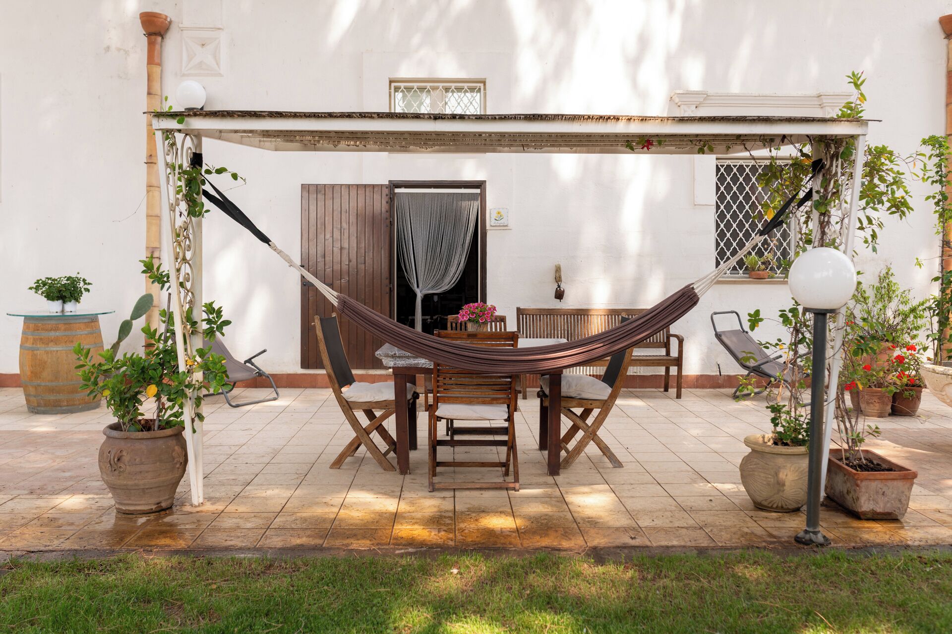 Una hamaca instalada sobre una mesa y sillas en un patio, con plantas en macetas y una pared blanca al fondo.
