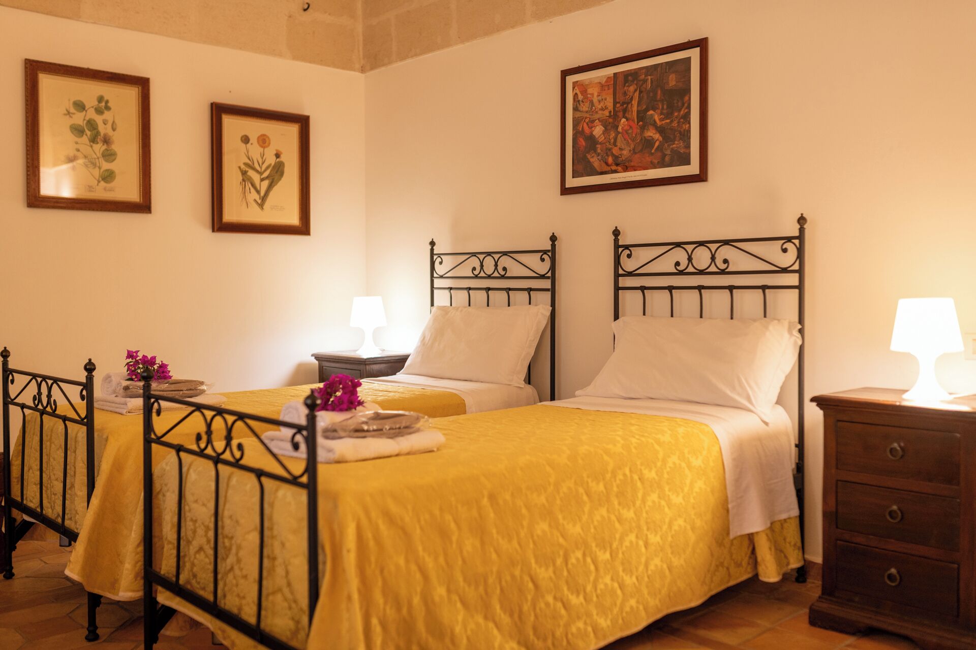 Habitación de hotel con dos camas, una mesita de noche, una lámpara e impresiones botánicas enmarcadas en la pared.