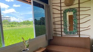 A room with a large window, a sofa, a small table with a plant, and a decorative wall panel. - Alamanda Family Villas & Pool (Sleman)