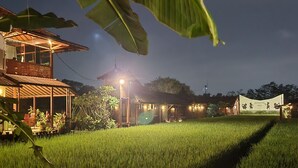 A well-lit, two-story building with a covered outdoor seating area and a rice field in the foreground. - Alamanda Family Villas & Pool (Sleman)