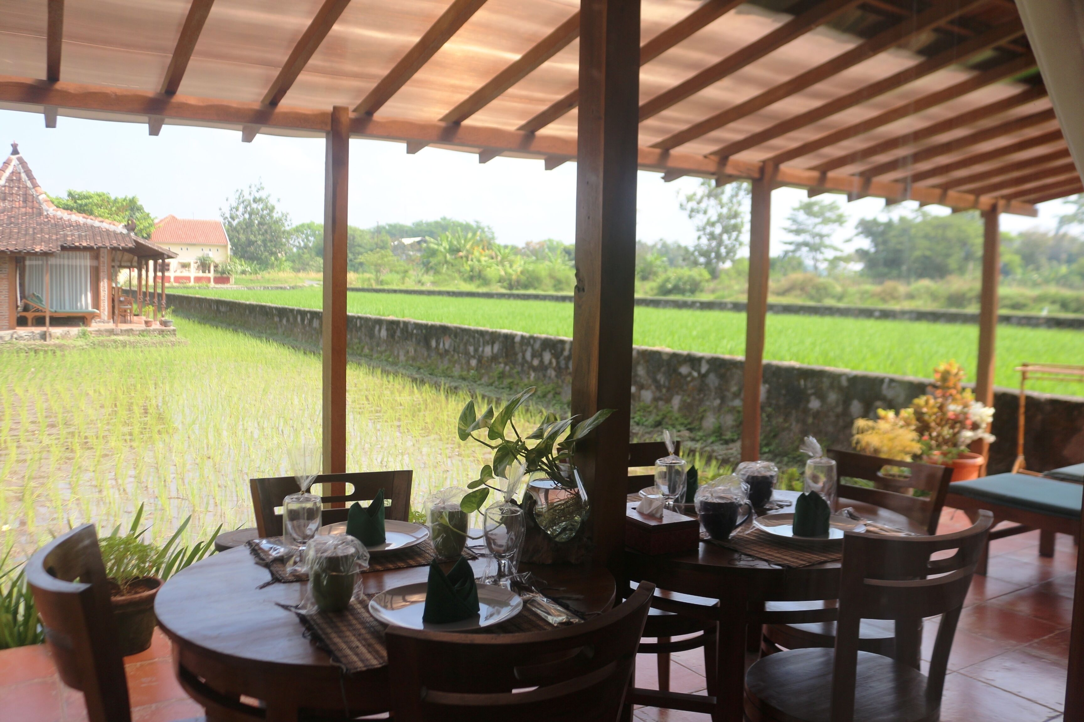 Uma área de refeições coberta com mesas preparadas para dois, com vista para um campo de arroz.