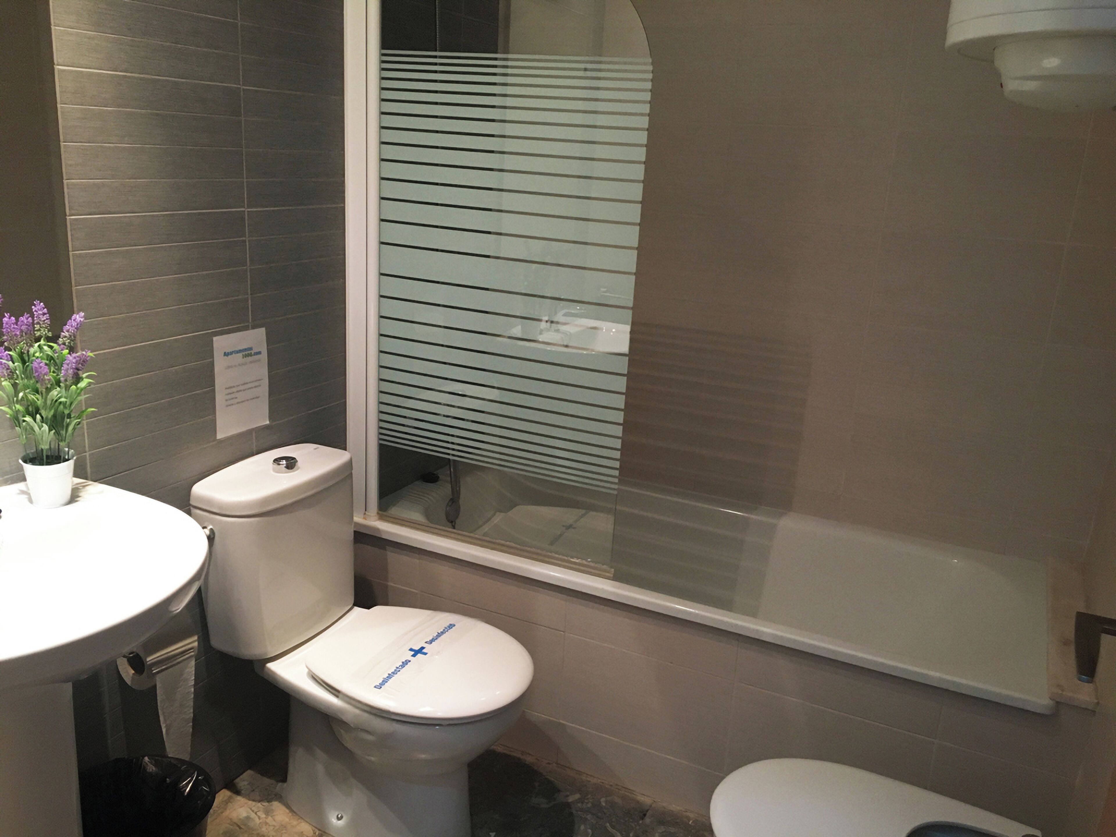 Une salle de bain avec des toilettes, un bidet et un lavabo. Il y a une fenêtre avec des stores, une plante en pot et un panneau accroché au mur.