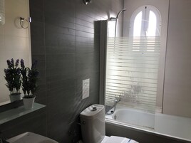 A bathroom with dark tiled walls, a white bathtub, and a window with blinds.