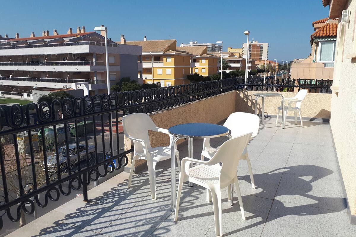 A terrace with white plastic chairs and a table, overlooking a residential area with buildings and a clear sky.
