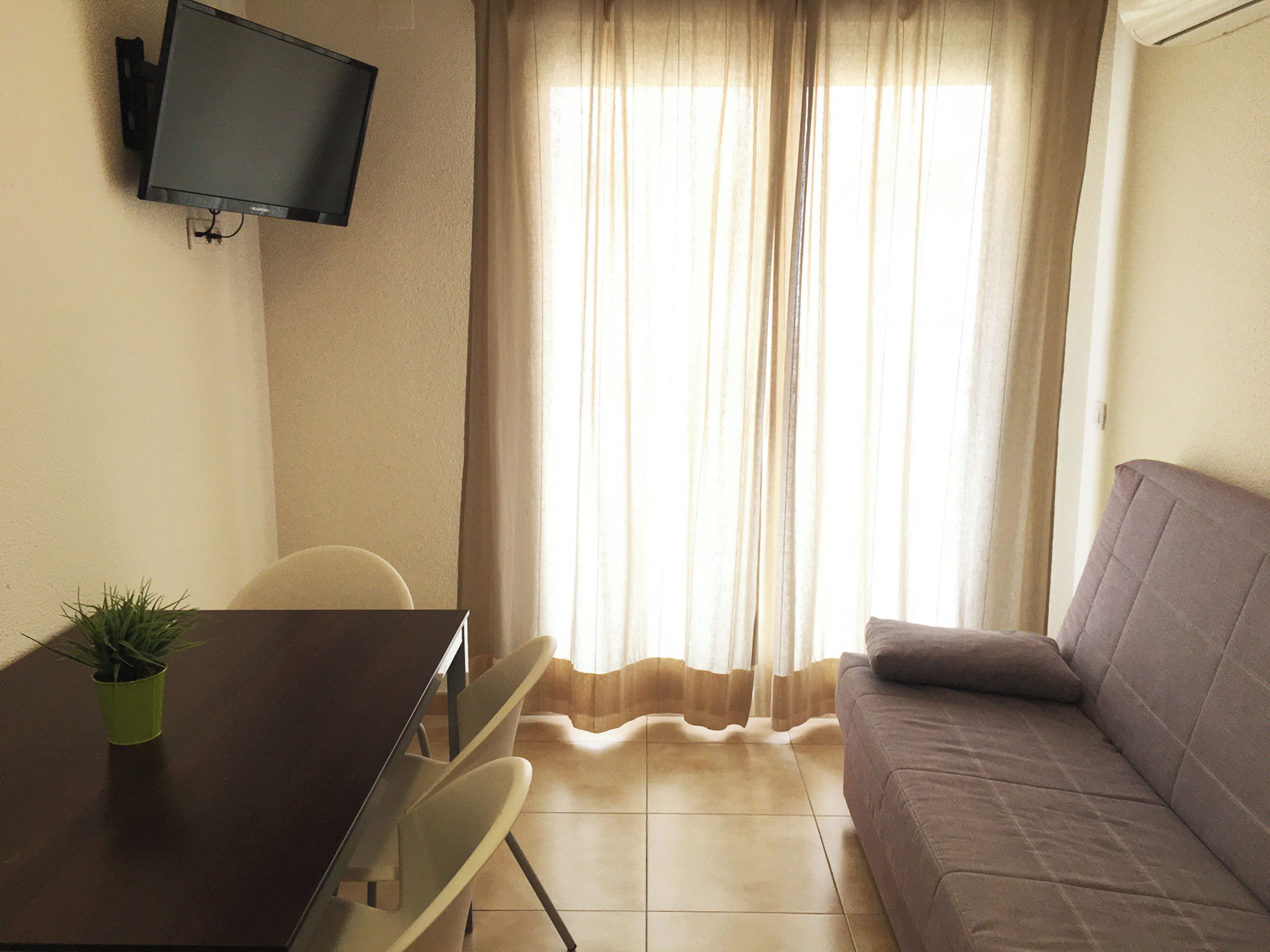 A small living area with a sofa, a TV mounted on the wall, a dining table with a plant, and a window with curtains.
