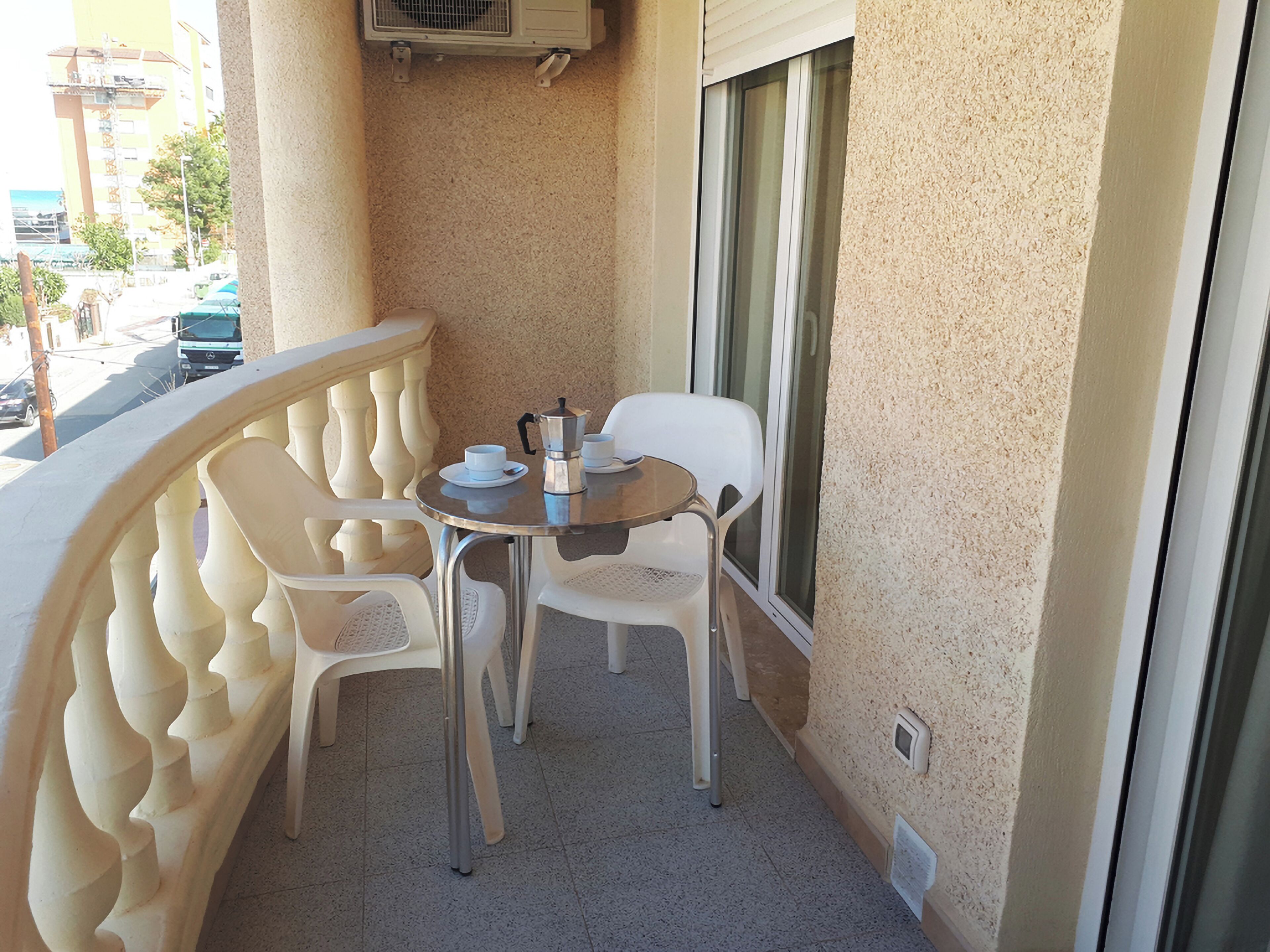 Un pequeño balcón con una mesa redonda y dos sillas blancas de plástico, una cafetera y una ventana con persianas.