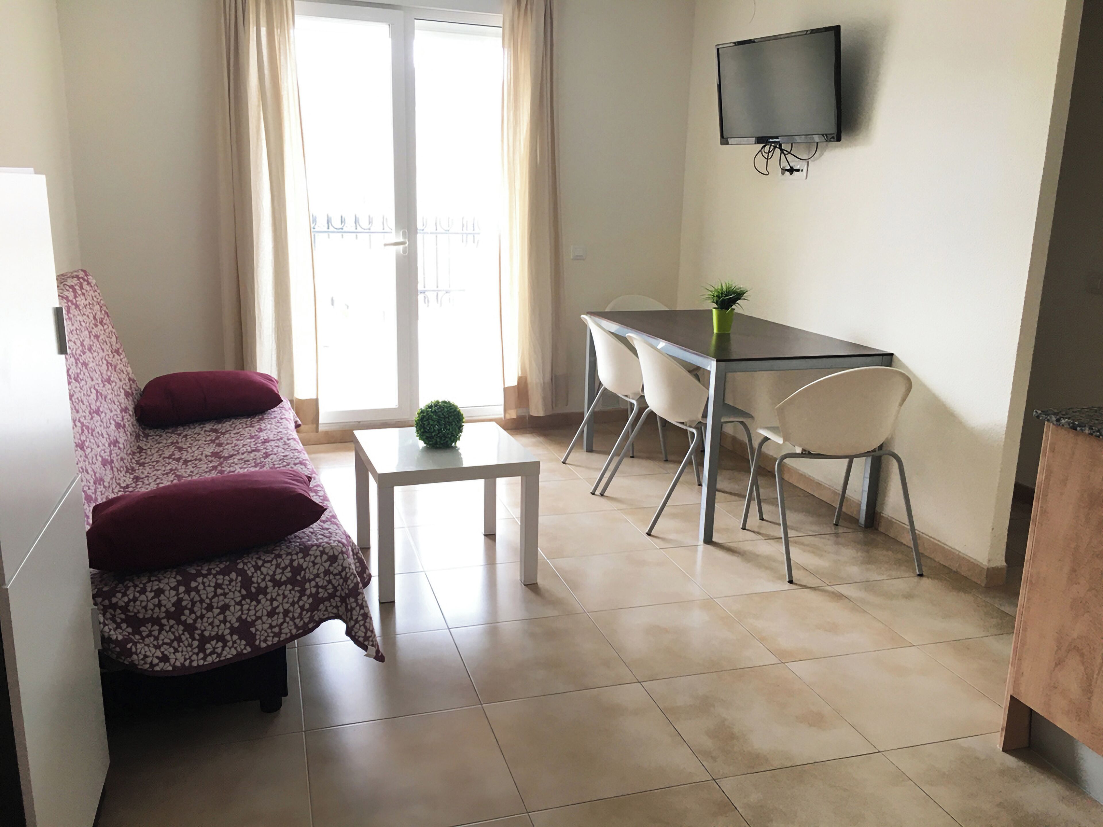 A compact living space with a sofa, a small table, a dining area with chairs, and a TV mounted on the wall.