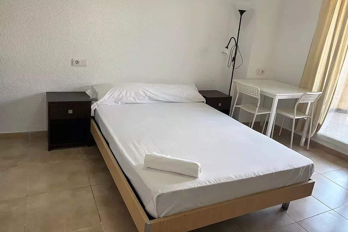 A single bed with a white mattress and pillows, a bedside table, a desk with a chair, and a floor lamp.