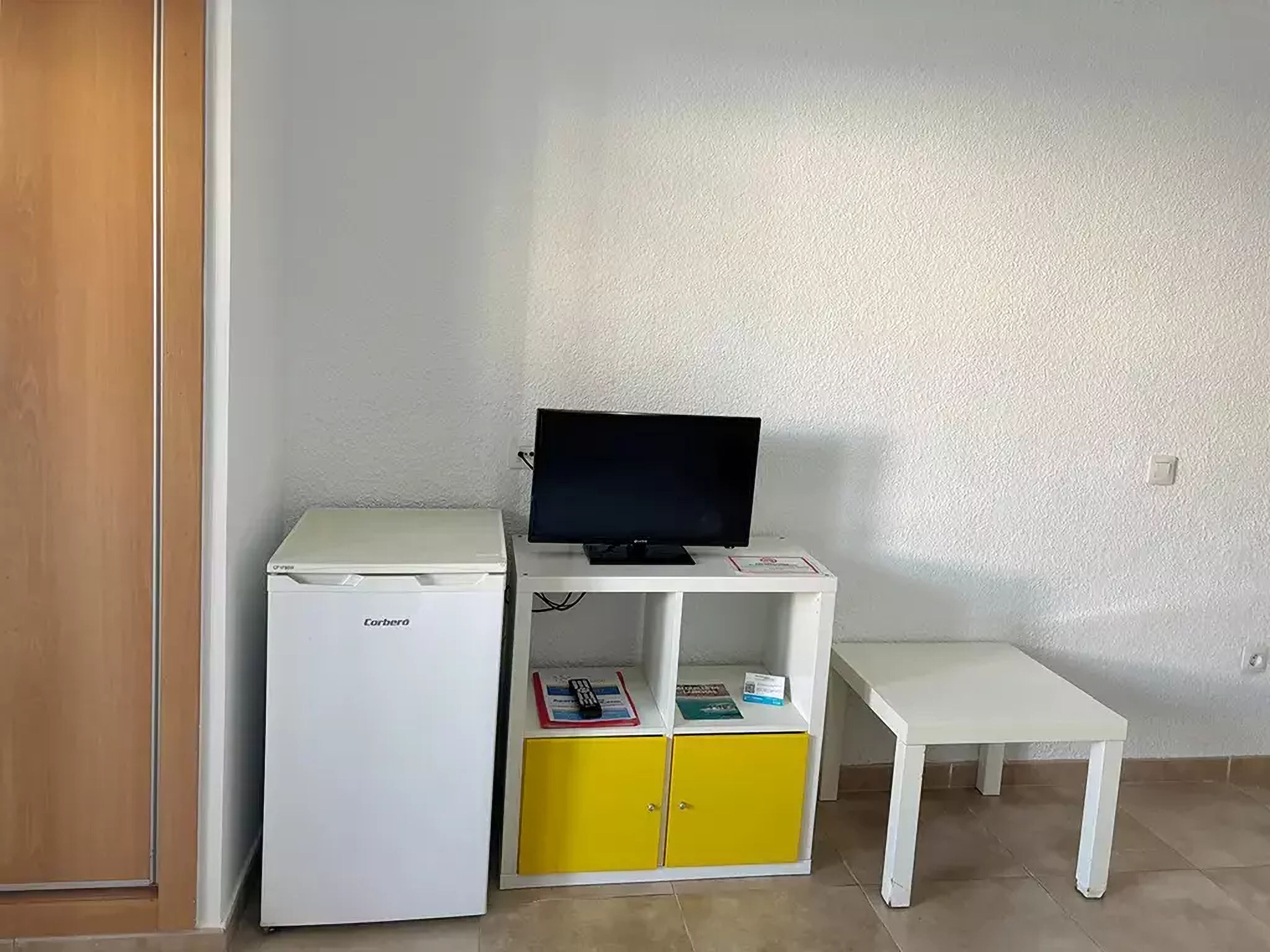 Habitación compacta con escritorio blanco, pequeño refrigerador blanco y unidad de almacenamiento amarilla con un televisor encima.