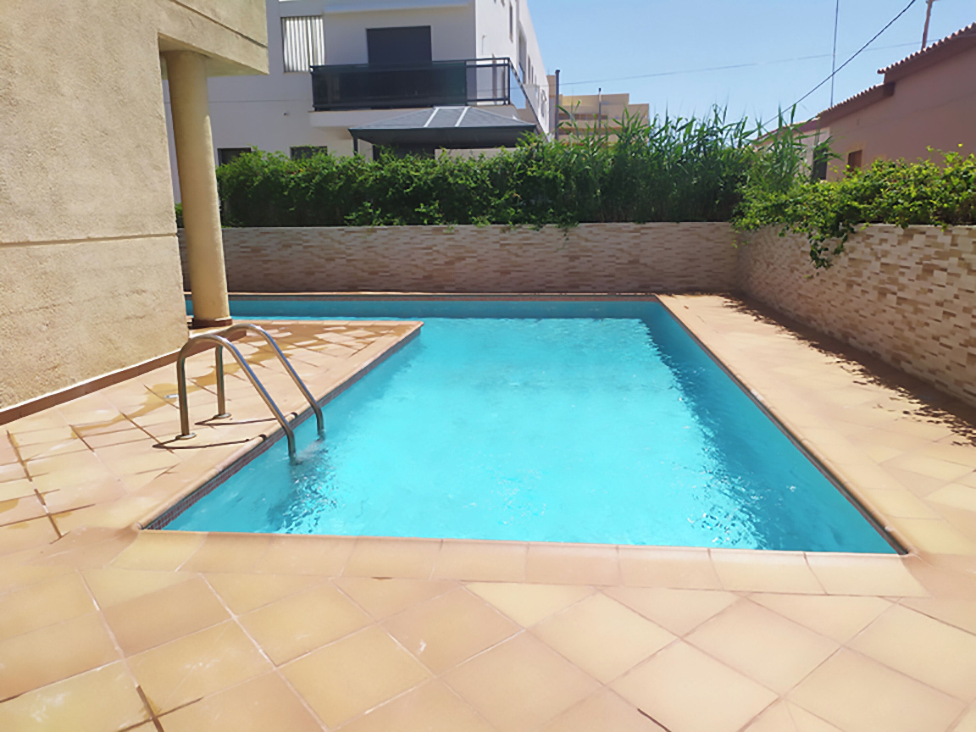 A rectangular swimming pool with a tiled deck, surrounded by a low stone wall and greenery.