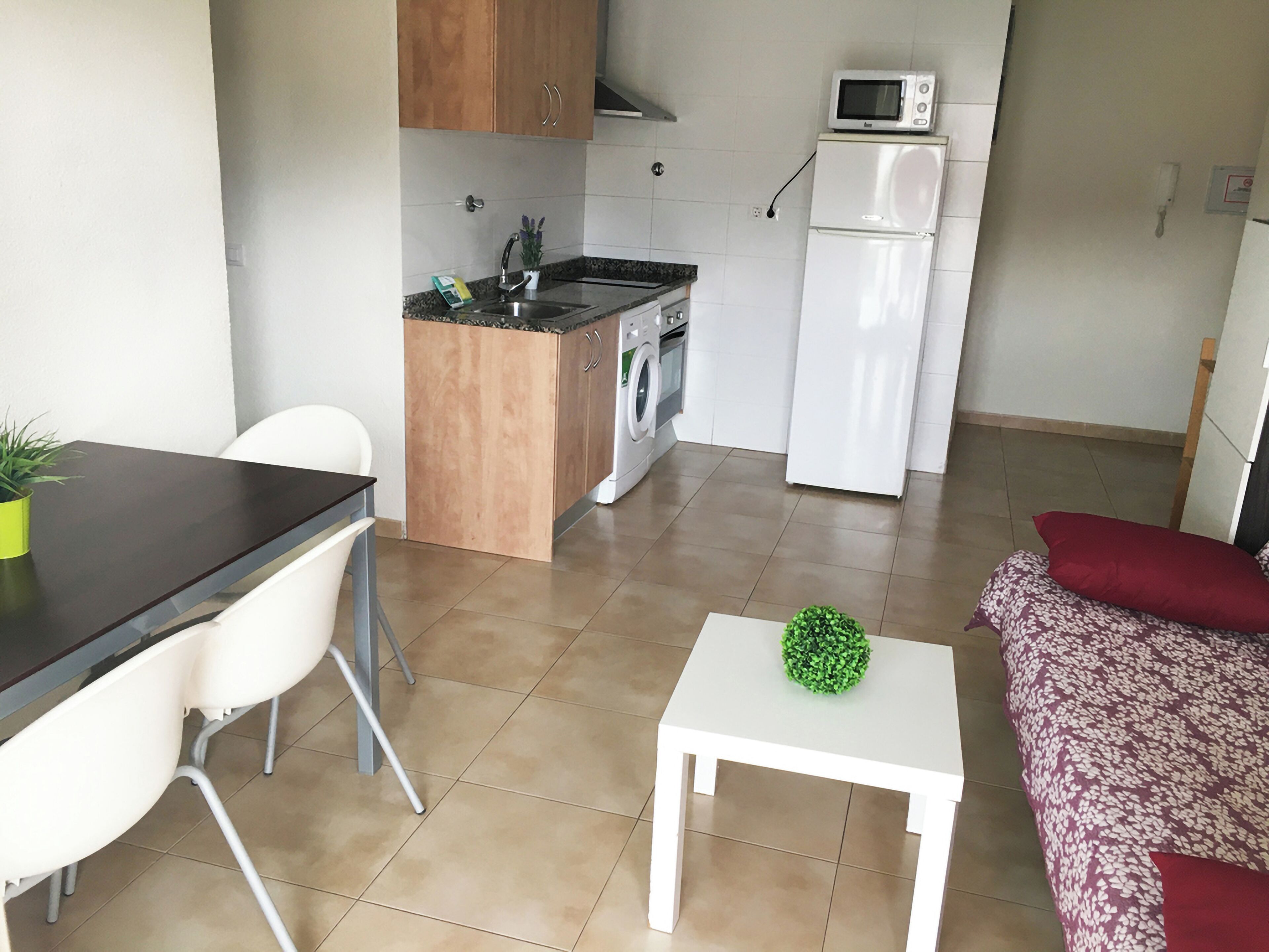 Eine kompakte Küche mit Mikrowelle, Backofen und Spüle. Es gibt einen Essbereich mit Tisch und Stühlen. Im Wohnbereich steht ein kleiner weißer Tisch mit einer grünen Pflanze.