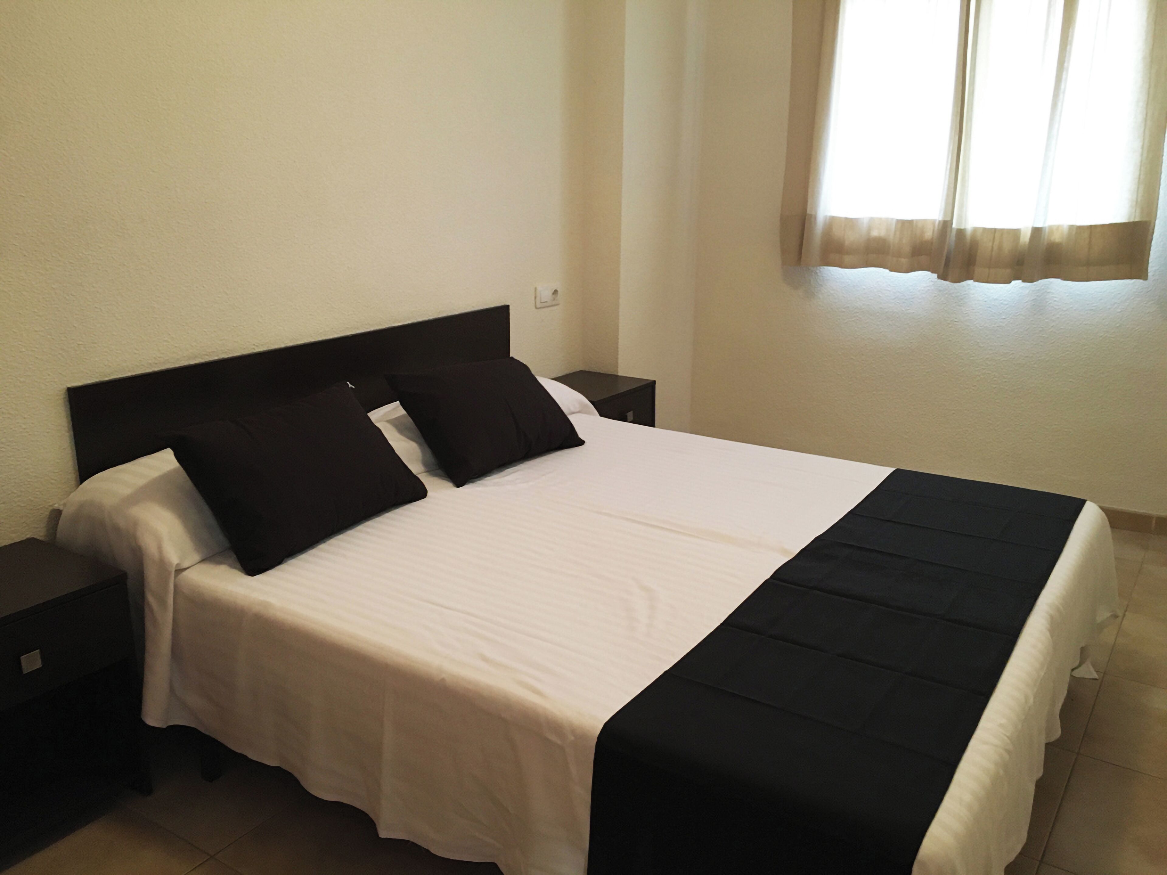 Una cama bien hecha con cabecera negra y ropa de cama blanca, una mesita de noche y una ventana con cortinas.
