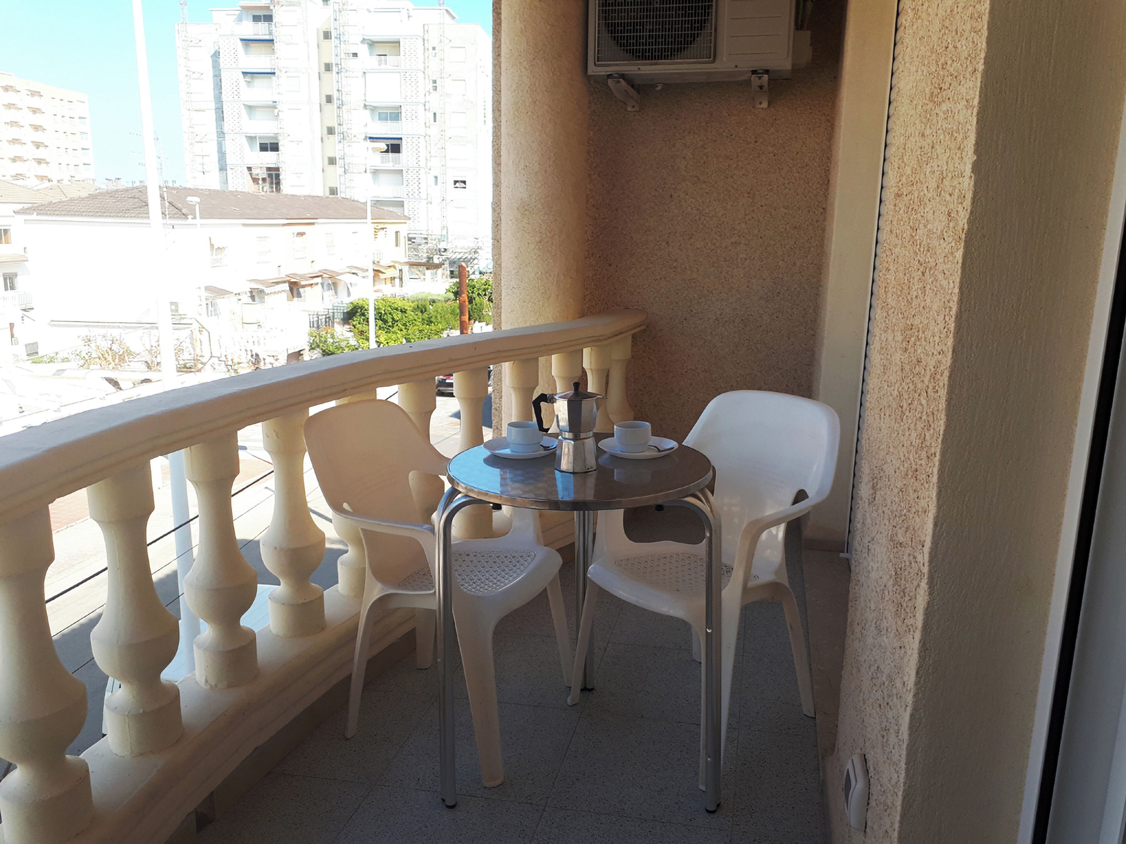 Een balkon met een ronde tafel en twee witte plastic stoelen, een airconditioning unit erboven, en een uitzicht op gebouwen op de achtergrond.