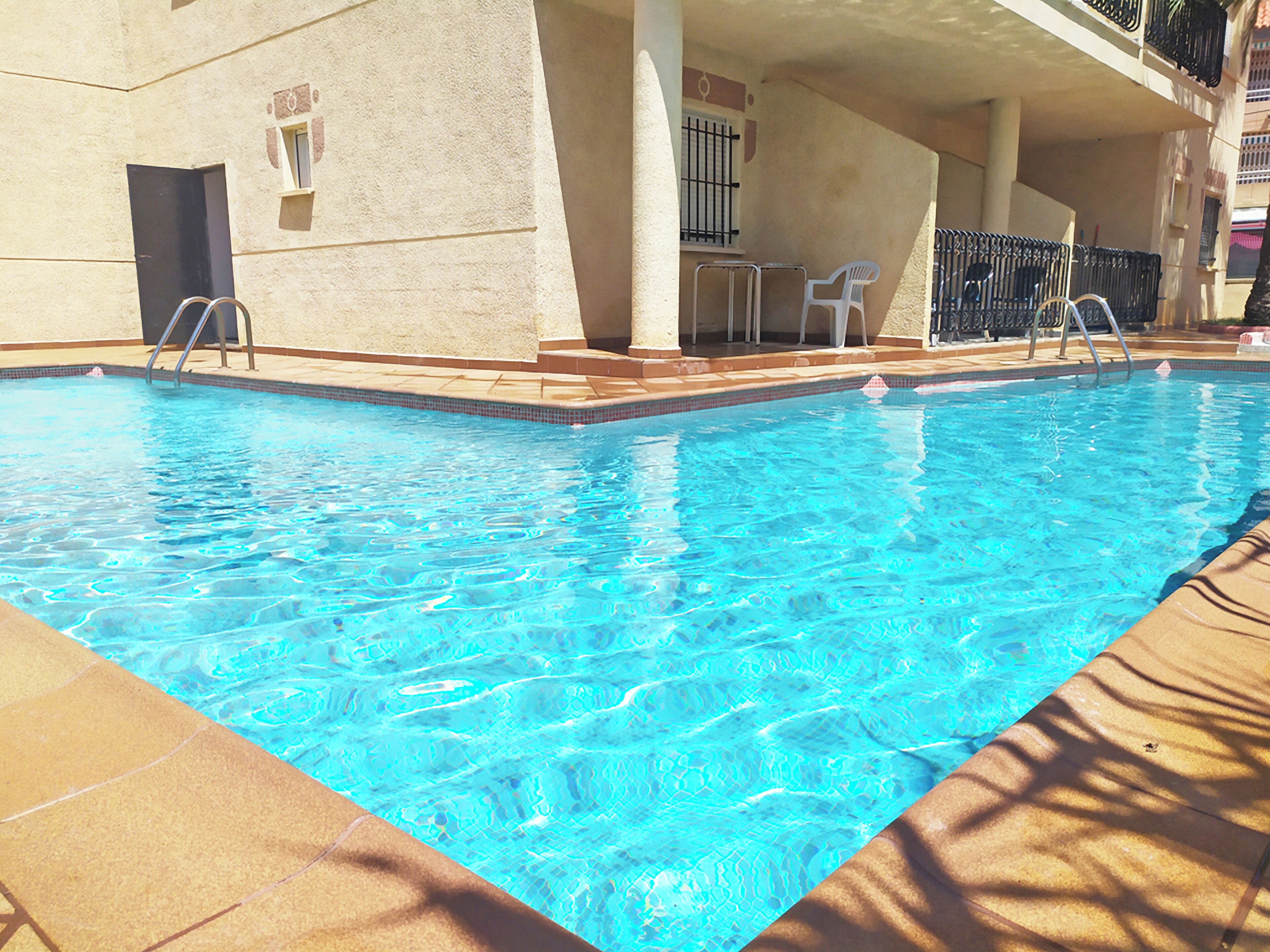 A swimming pool with clear blue water, surrounded by a tiled deck and a building with a beige facade.