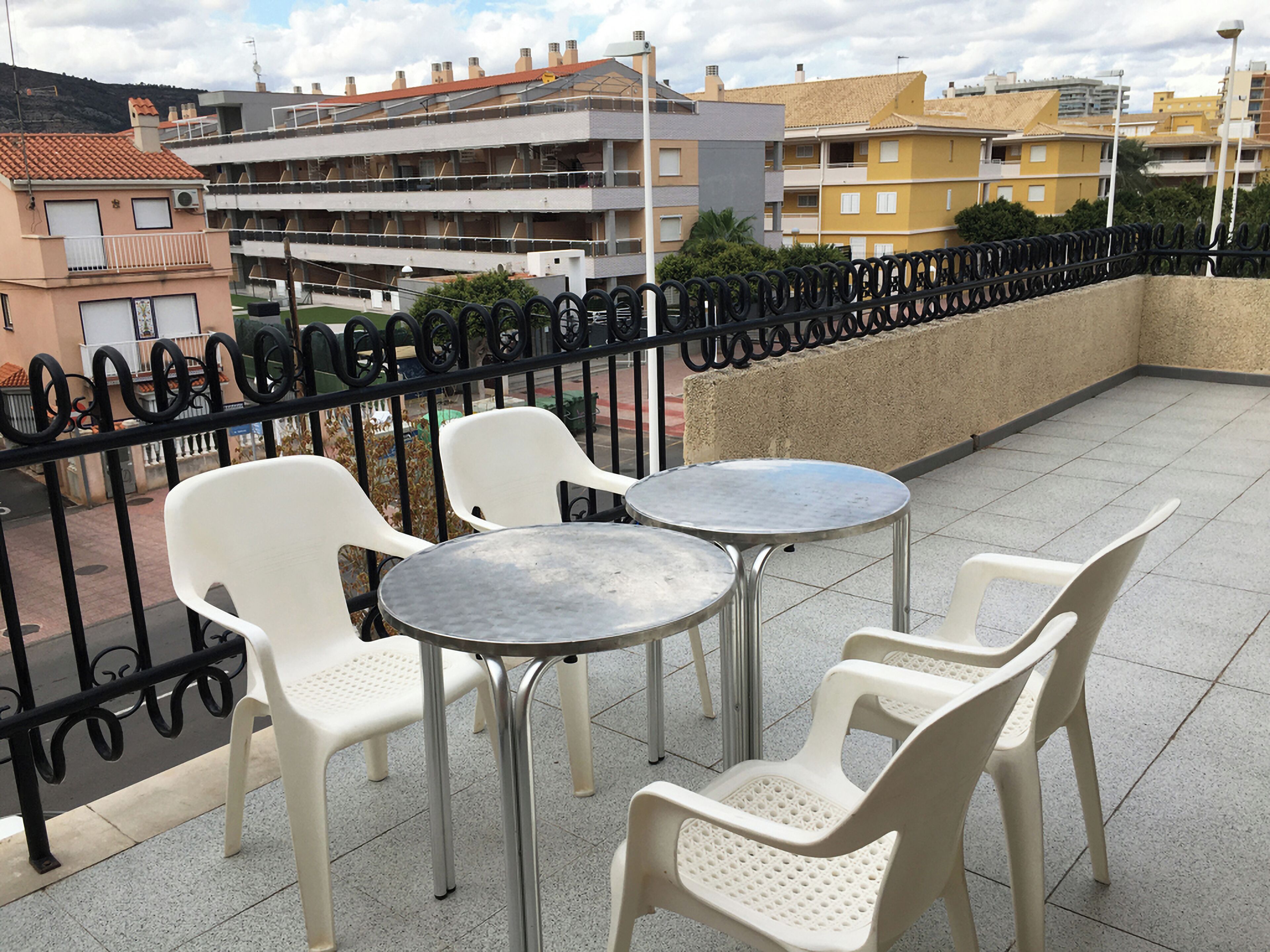 An outdoor terrace with white plastic chairs and metal tables, surrounded by a black wrought iron fence.
