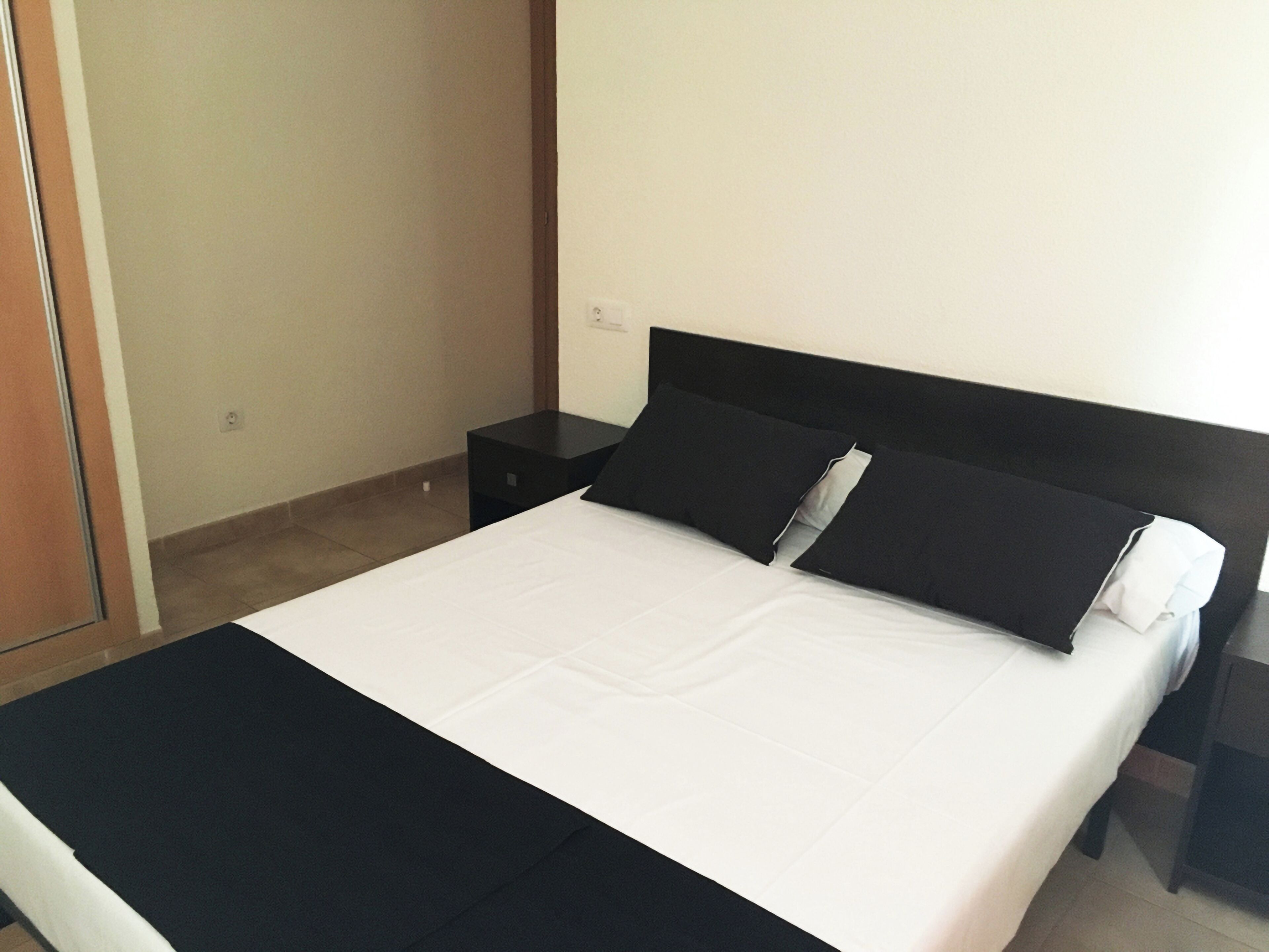 Una cama bien hecha con cabecera negra y ropa de cama blanca, una mesita de noche pequeña y una pared lisa con una puerta a la izquierda.