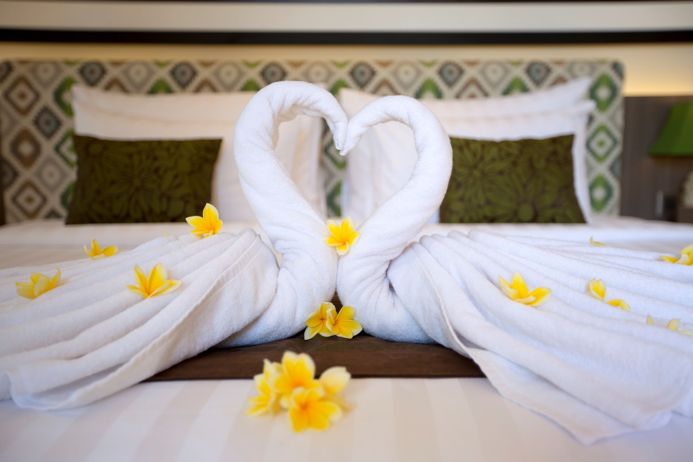 A bed with white towels arranged in a heart shape, yellow flowers scattered on the bed, and a patterned headboard.