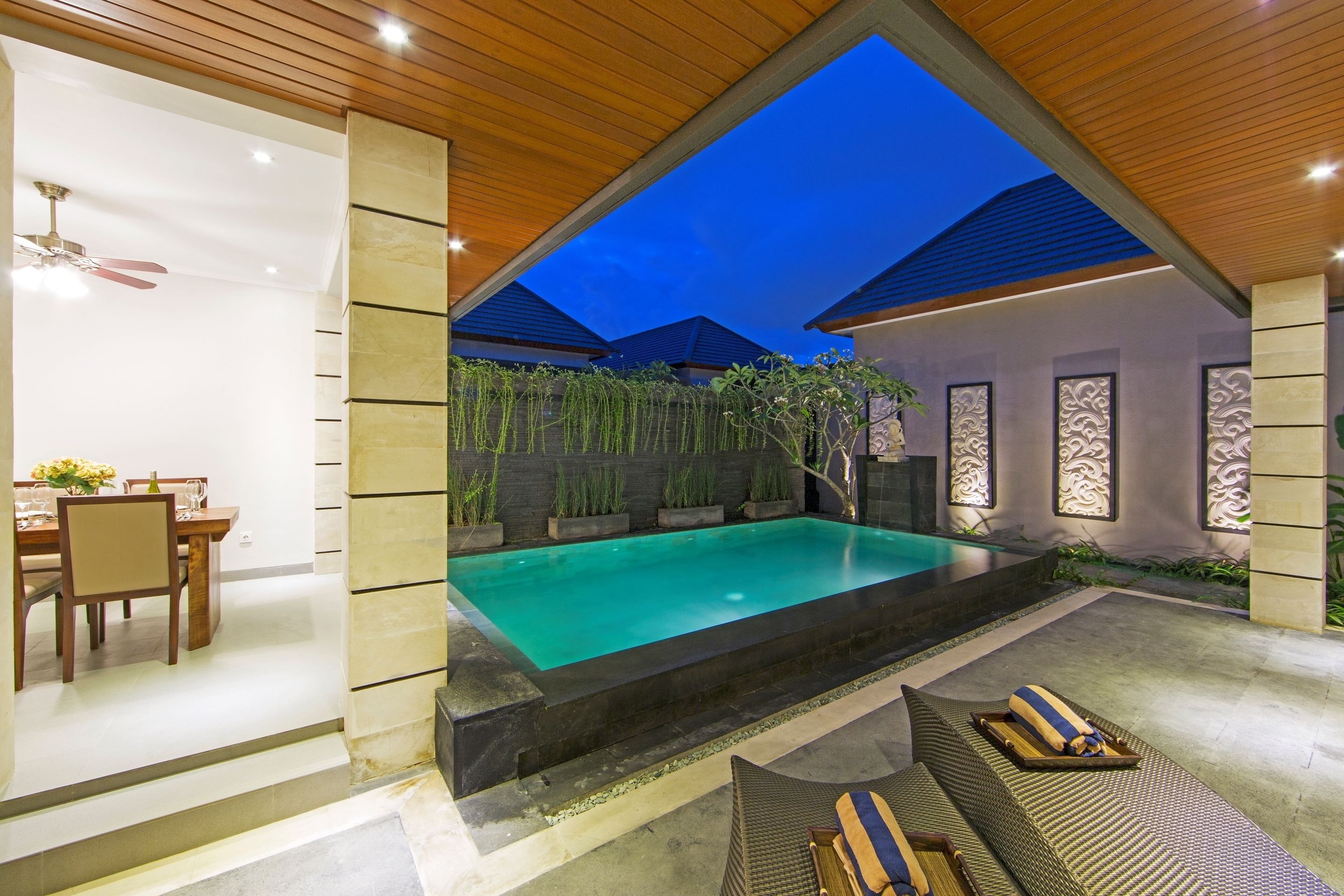 A modern outdoor pool area with a wooden ceiling, a dining setup, and a lounge area with cushions.