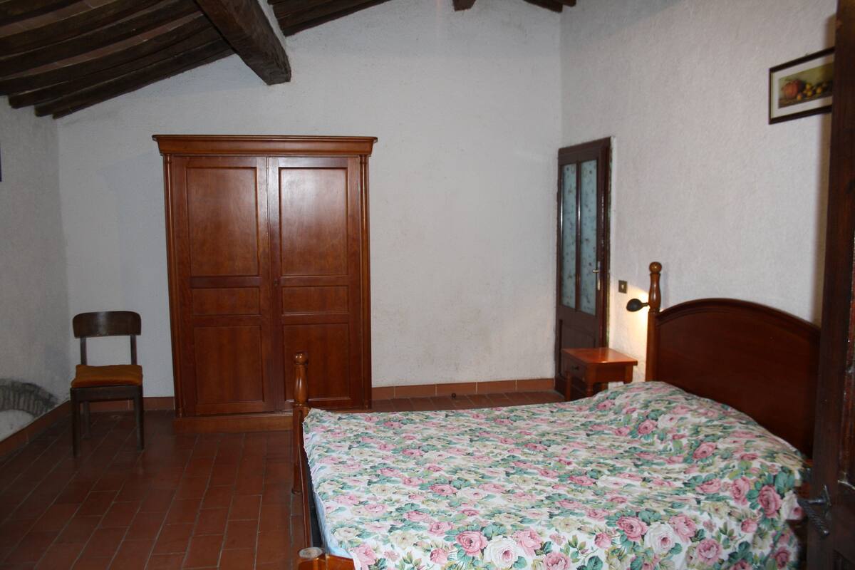 Une chambre à coucher avec une armoire en bois, un lit recouvert d’une couvre-lit à motifs floraux, une chaise et une petite table de chevet.