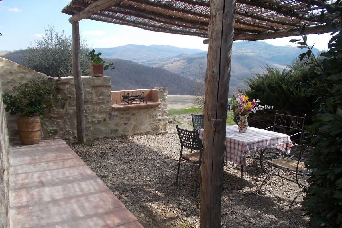 Une terrasse extérieure avec un banc en pierre, une table recouverte d’une nappe à carreaux et une pergola rustique.