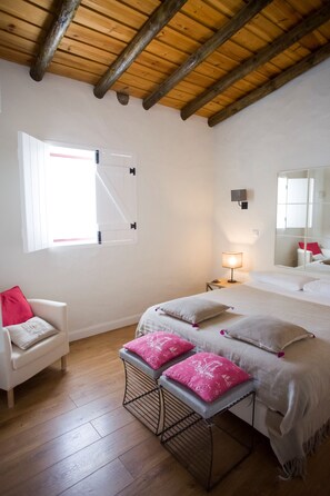 A bedroom with a wooden ceiling, a bed with pink and white bedding, a white chair with a red pillow, a bench with pink cushions, a bedside table with a lamp, and a large mirror. - Herdade dos Castelejos (Castro Verde)