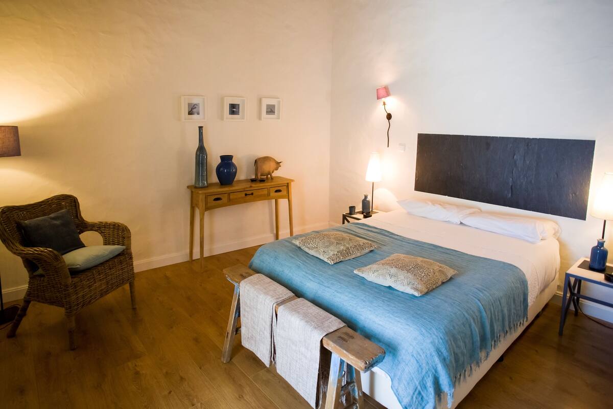 A bedroom with a large bed, a wooden bench, a wicker chair, a small table with a vase, and a wall-mounted lamp.