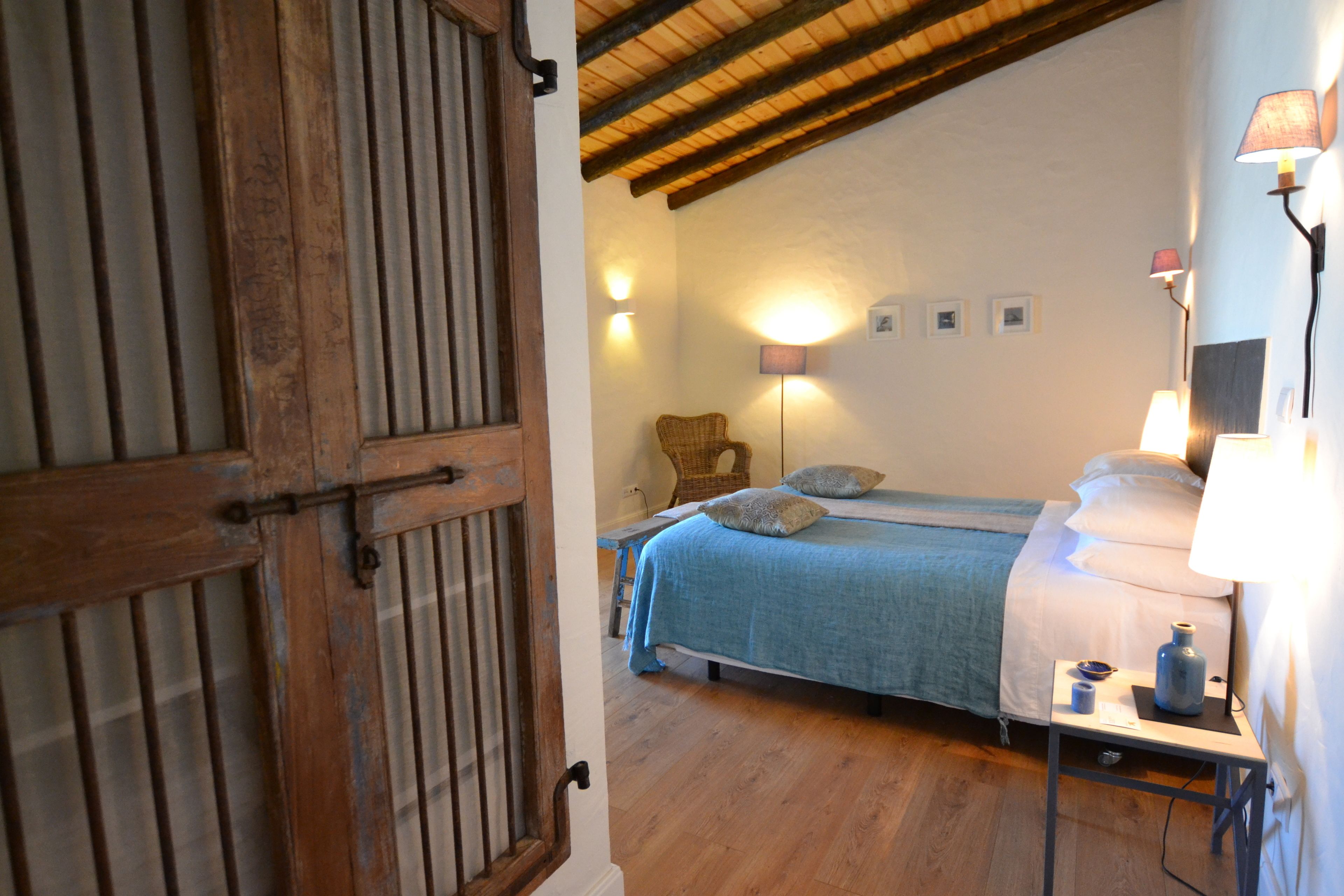 A bedroom with a wooden door, a bed with a blue blanket, a chair, and wall-mounted lamps.