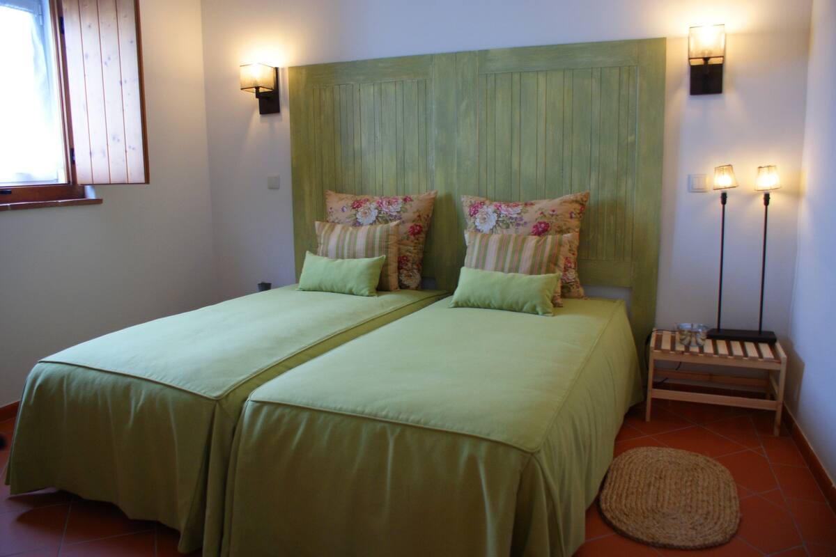 A room with two beds, a headboard, wall-mounted lights, a bedside table, and a window.