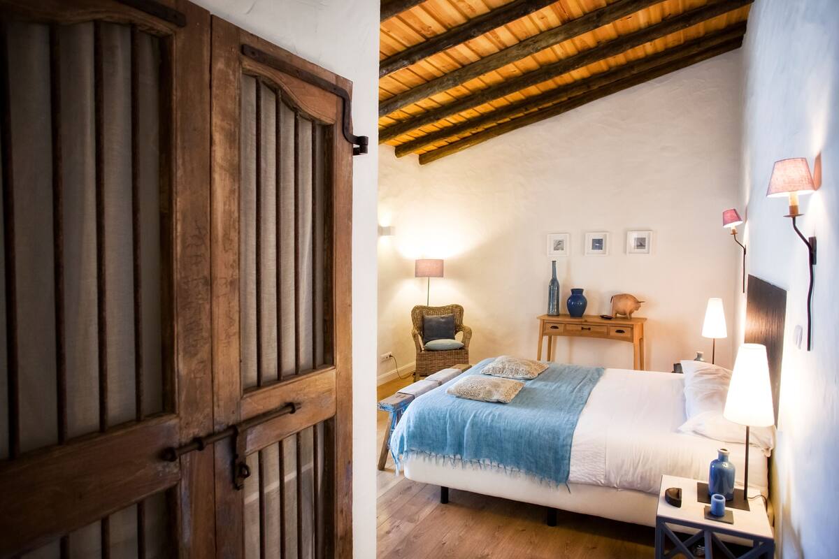 A bedroom with a wooden door, a bed with a blue blanket, a wicker chair, and two bedside tables with lamps.