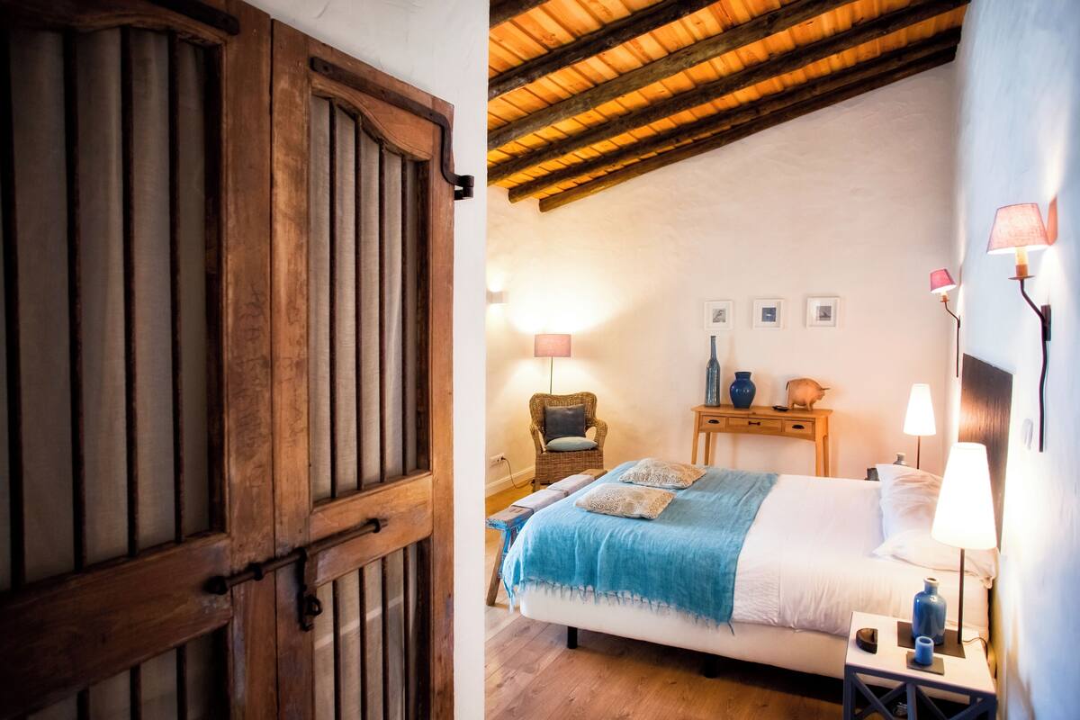 A bedroom with a wooden door, a bed with a blue blanket, a wicker chair, and two bedside tables with lamps.