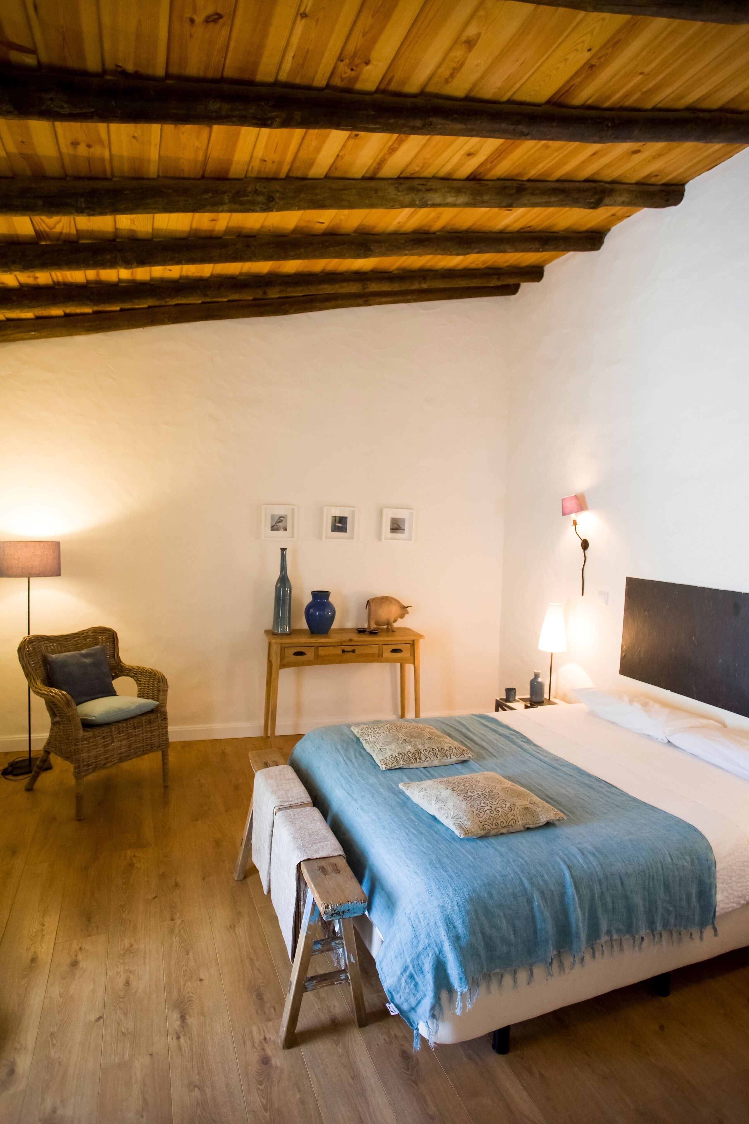 Una camera da letto con soffitto in legno, un letto con coperta blu, un comodino con una lampada e una sedia.