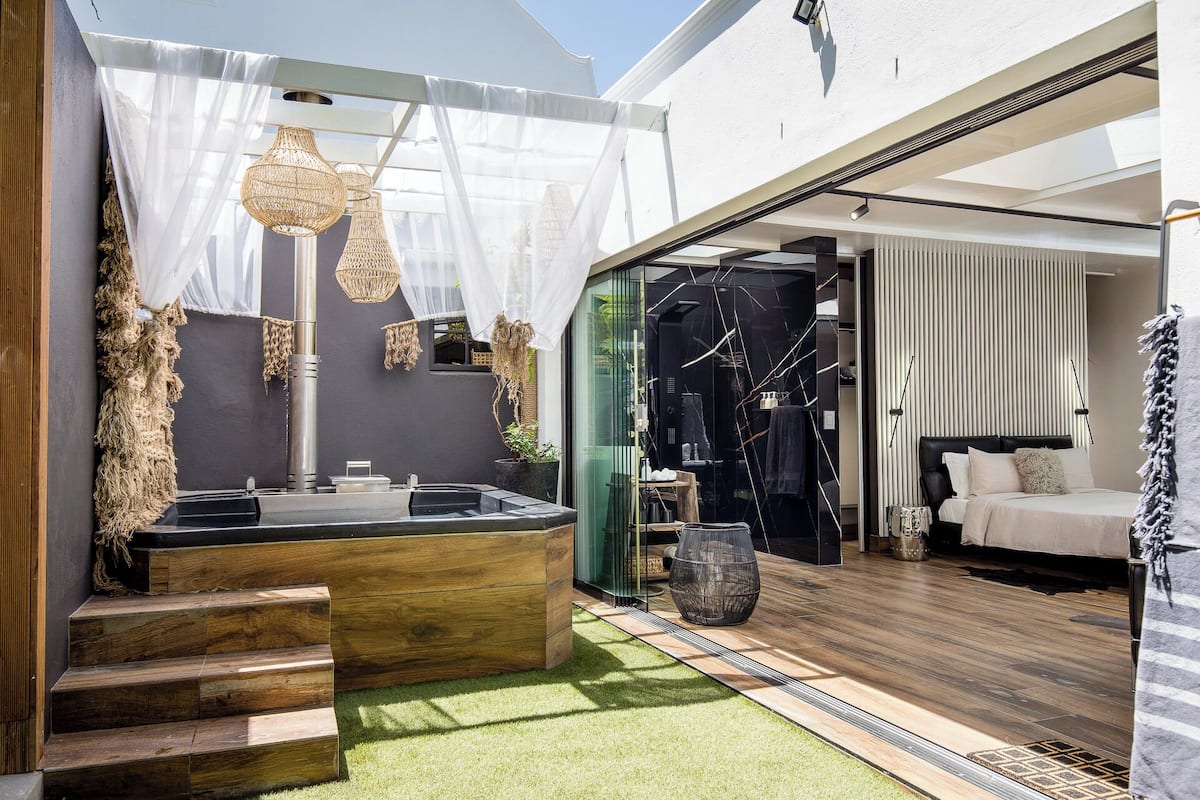 A modern outdoor bathroom with a wooden bench, a hot tub, and a glass-enclosed shower area.