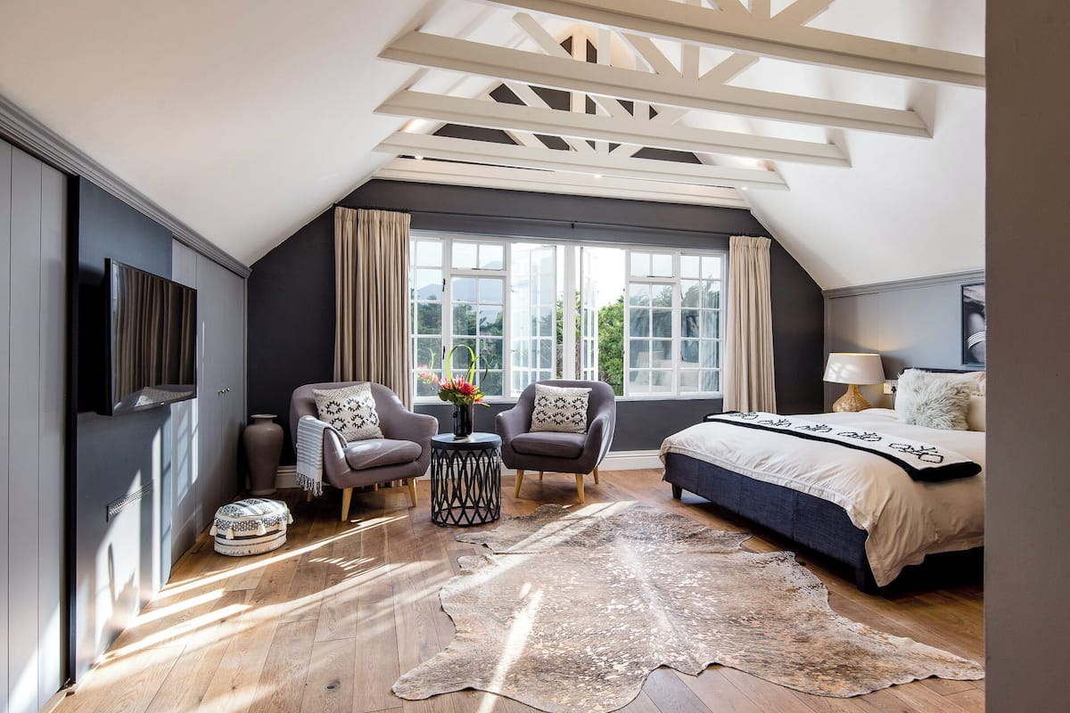 A modern bedroom with a sloped ceiling, a large bed, two armchairs, a small table with a vase, and a large window with curtains.