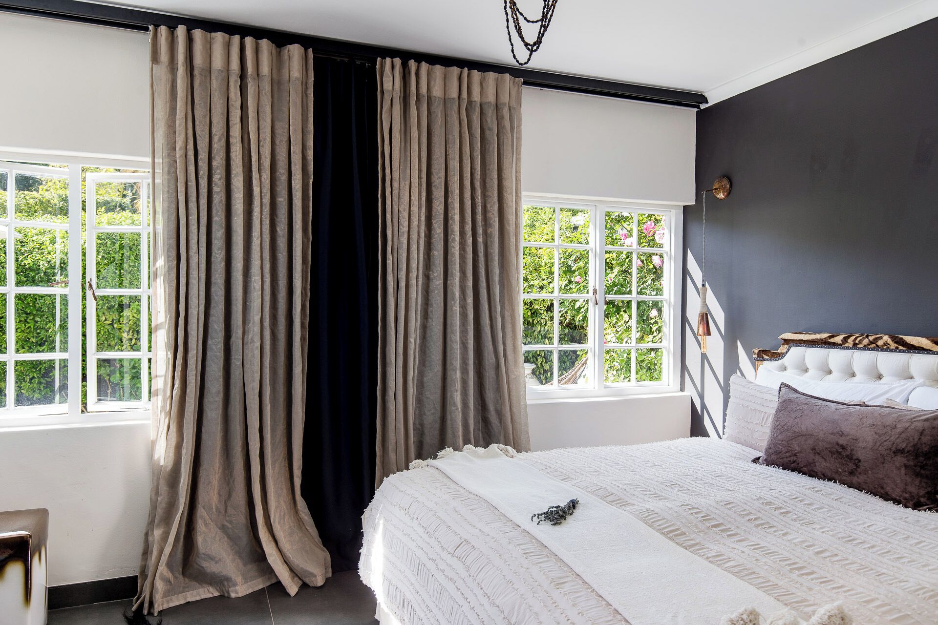 A bedroom with a large bed, two windows with curtains, a dark wall, and a headboard with a decorative element.