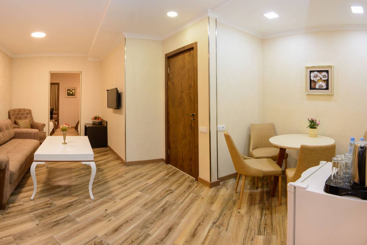 A modern hotel room with a wooden floor, a sofa, a coffee table, a dining table with chairs, a TV mounted on the wall, and a door.
