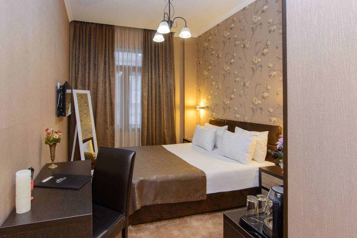 A hotel room with a bed, a desk with a laptop, a chair, a nightstand with a lamp, and a mirror on the wall.