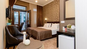 A hotel room with a bed, a desk, a chair, a small table, a TV, and a mini-fridge. - Hotel King David (Tbilisi)