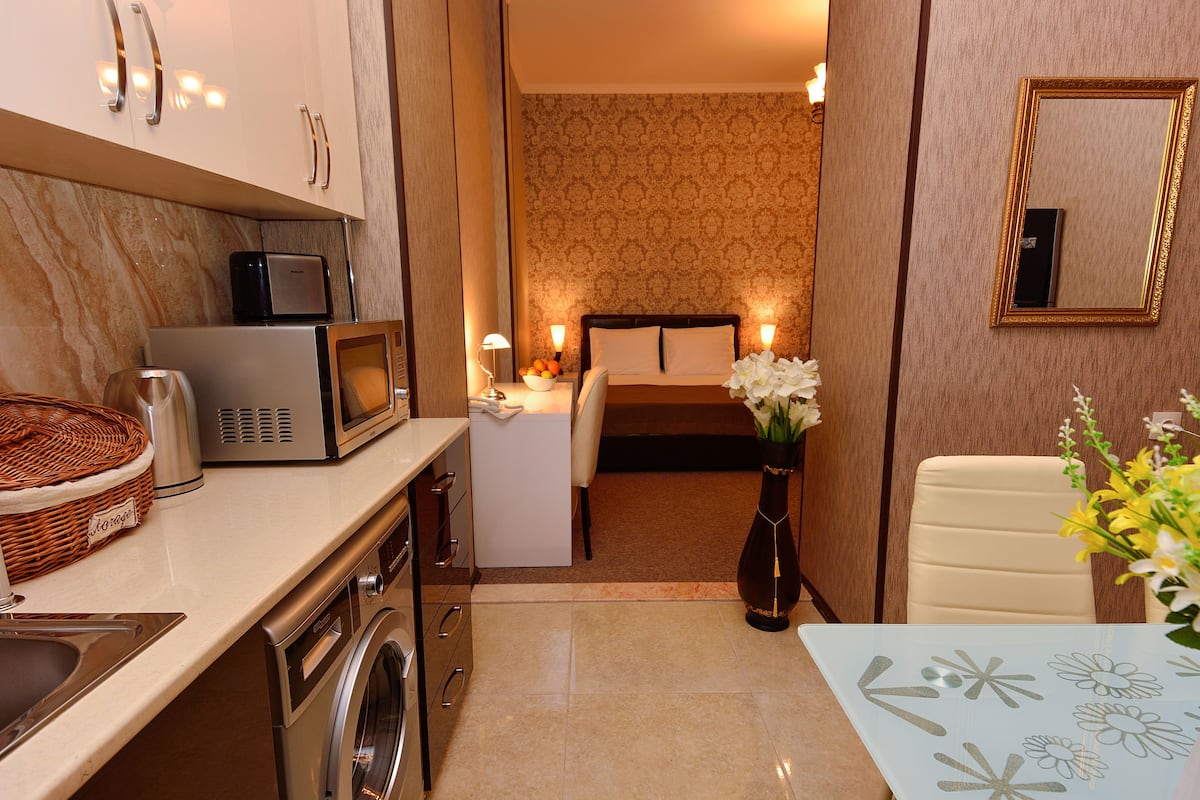 A compact hotel room with a kitchenette, a bed with a nightstand, a small table with a vase, and a mirror on the wall.