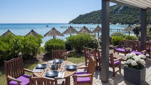 Ein überdachter Essbereich im Freien mit Holztischen und violetten Kissen, mit Blick auf einen Strand mit Reetdachschirmen und einem Steg.